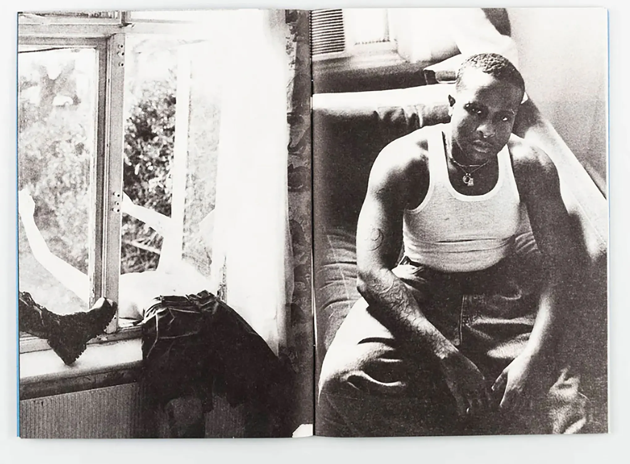 Black-and-white photo of a person with short hair sitting on a sofa, wearing a white vest and loose trousers. They have tattoos on one arm and are looking at the camera. Sunlight comes through a window with clothes draped on its sill beside them.
