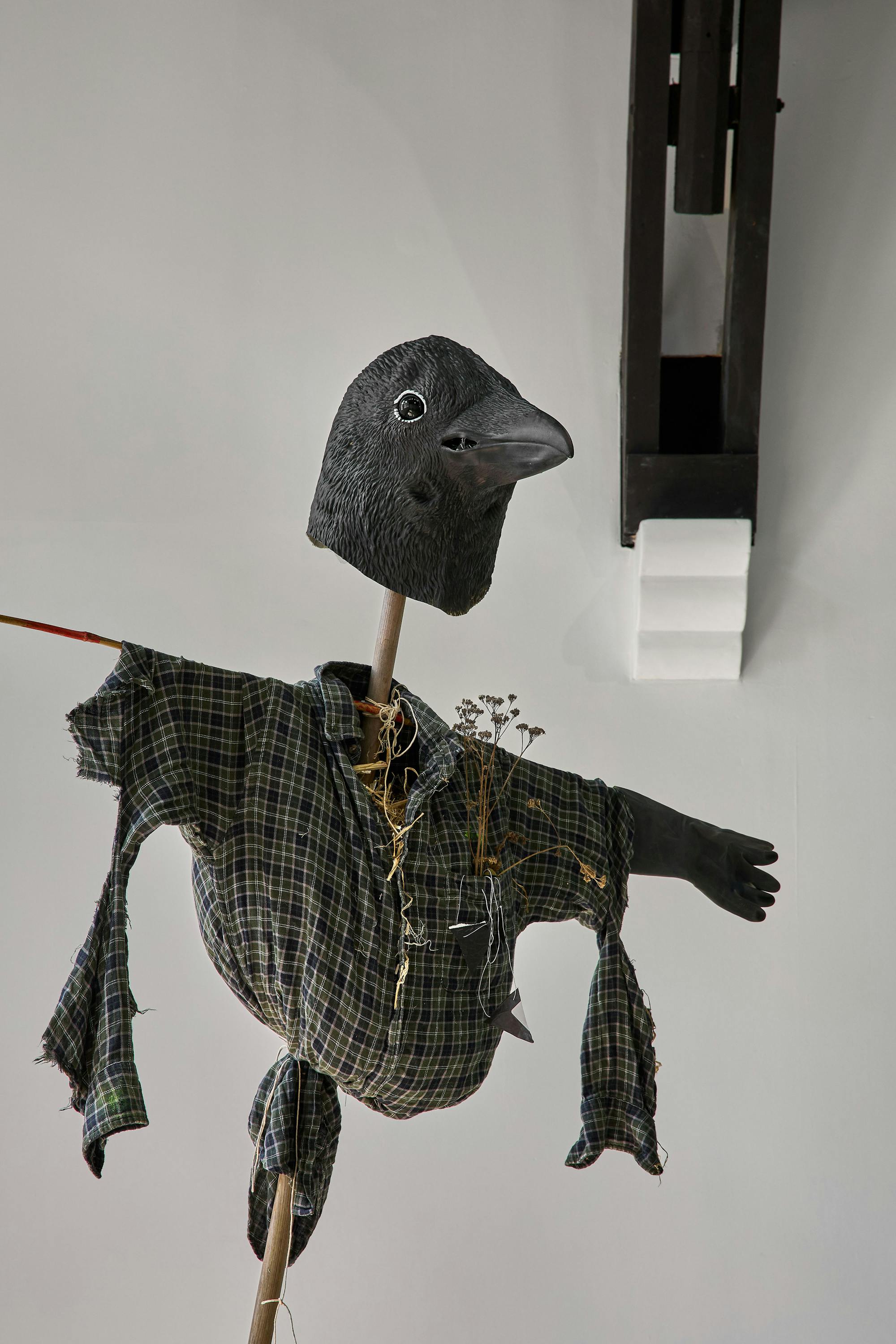 A scarecrow with a large black bird head, button eyes, and open beak wears a checked shirt stuffed with straw and dried flowers. Its arms are spread wide and the background shows a plain white ceiling with a wooden beam.