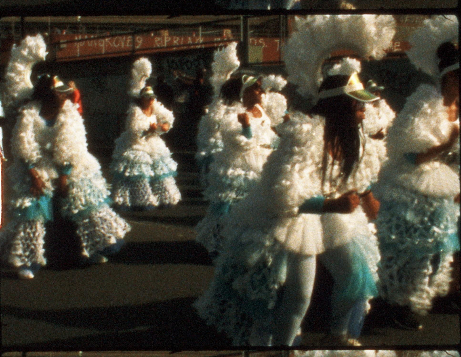 A group of people in elaborate, ruffled white and blue costumes and headdresses dance together on a street during a parade or festival. The background shows a faded wall with graffiti. The image is slightly blurred, suggesting motion and vibrant energy.