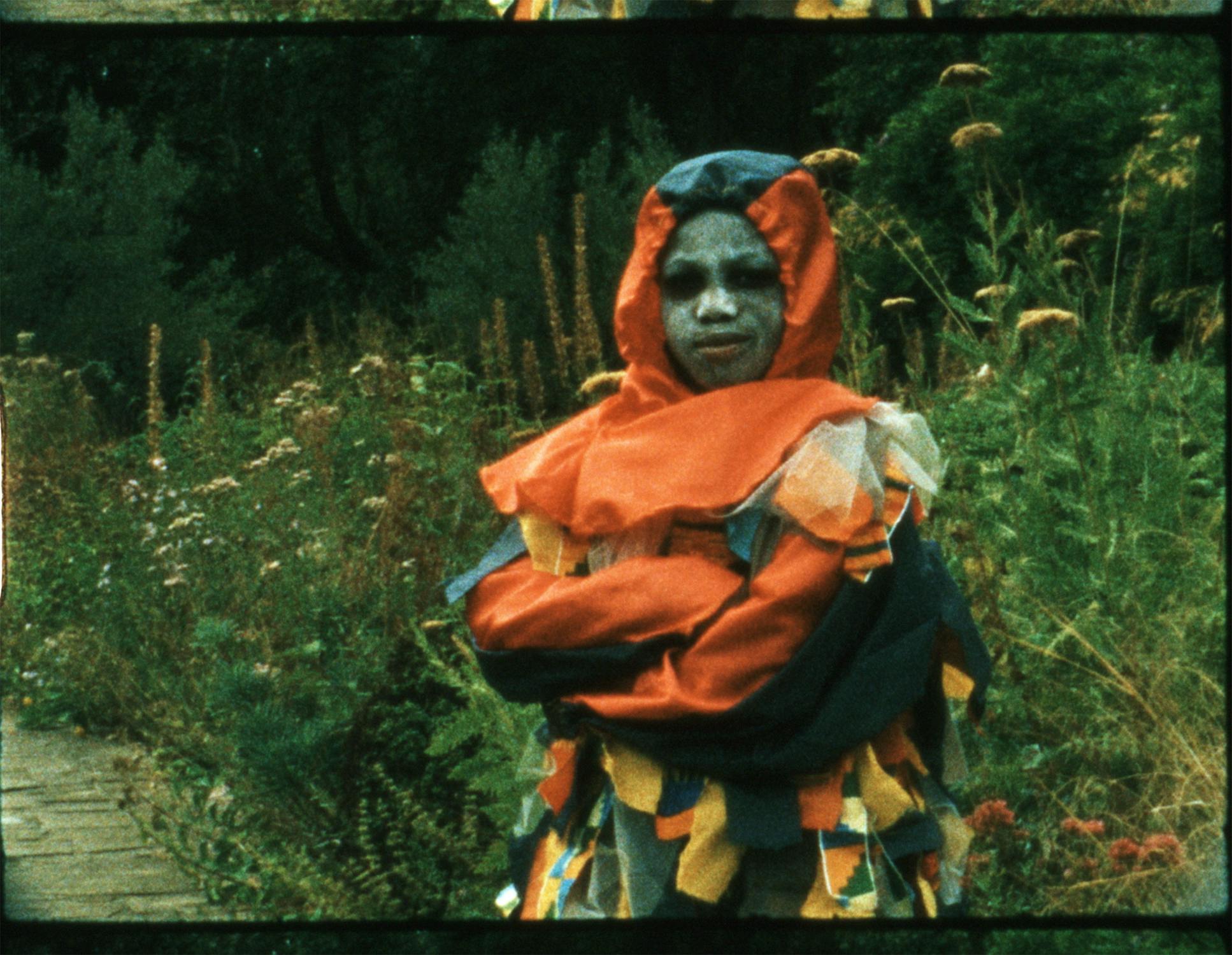 A person in a vibrant, patchwork costume with a red hood and colourful fabric strips stands outdoors on a path, arms crossed. Their face is painted pale bluish-grey, with dark eye make-up. Lush greenery and wildflowers fill the background.