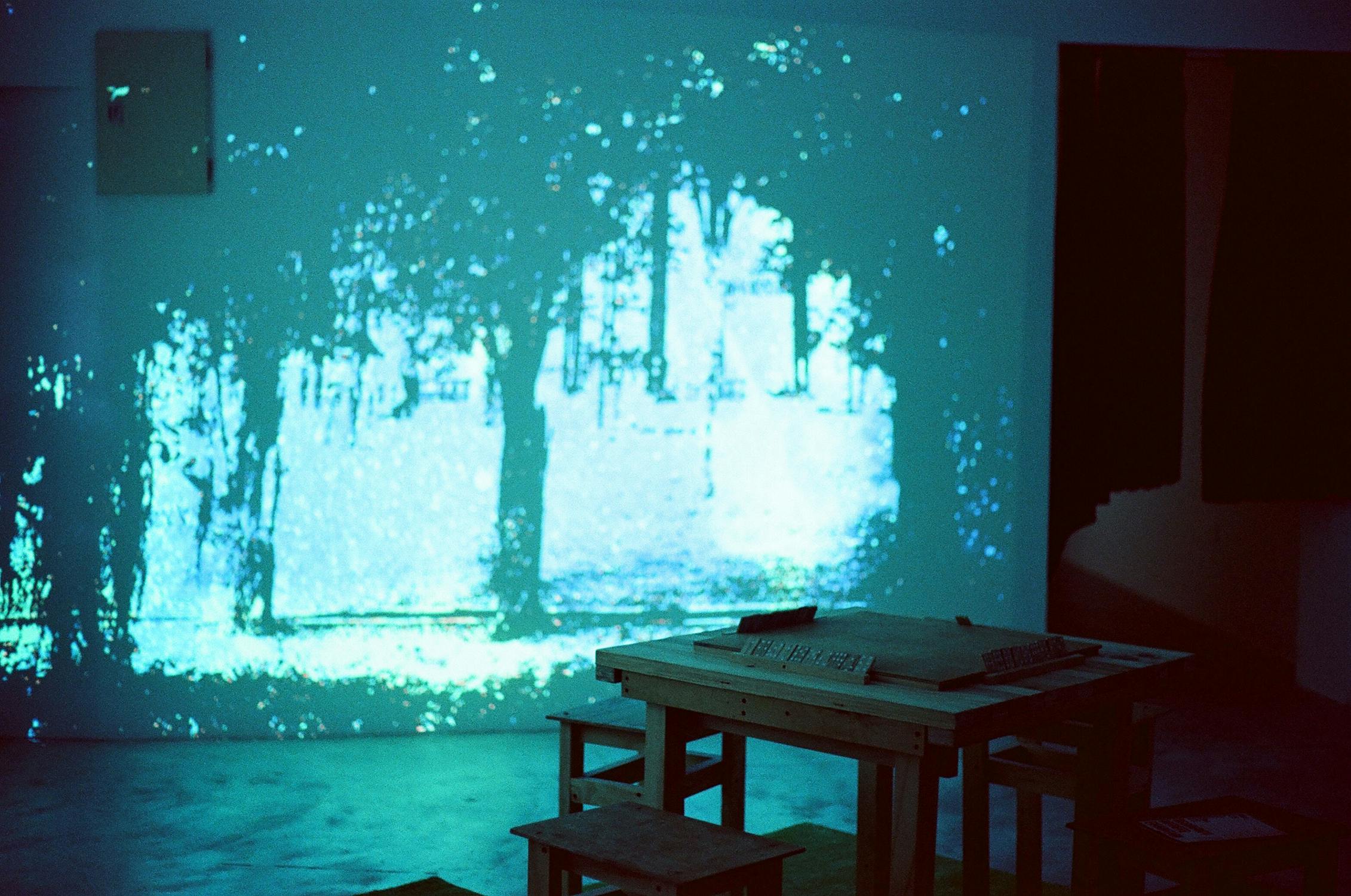 A dimly lit room features a projection of a blue-tinted forest scene on the wall. In the foreground, there’s a wooden table with stools, partially illuminated by the light from the projection, creating an atmospheric, dreamy effect.