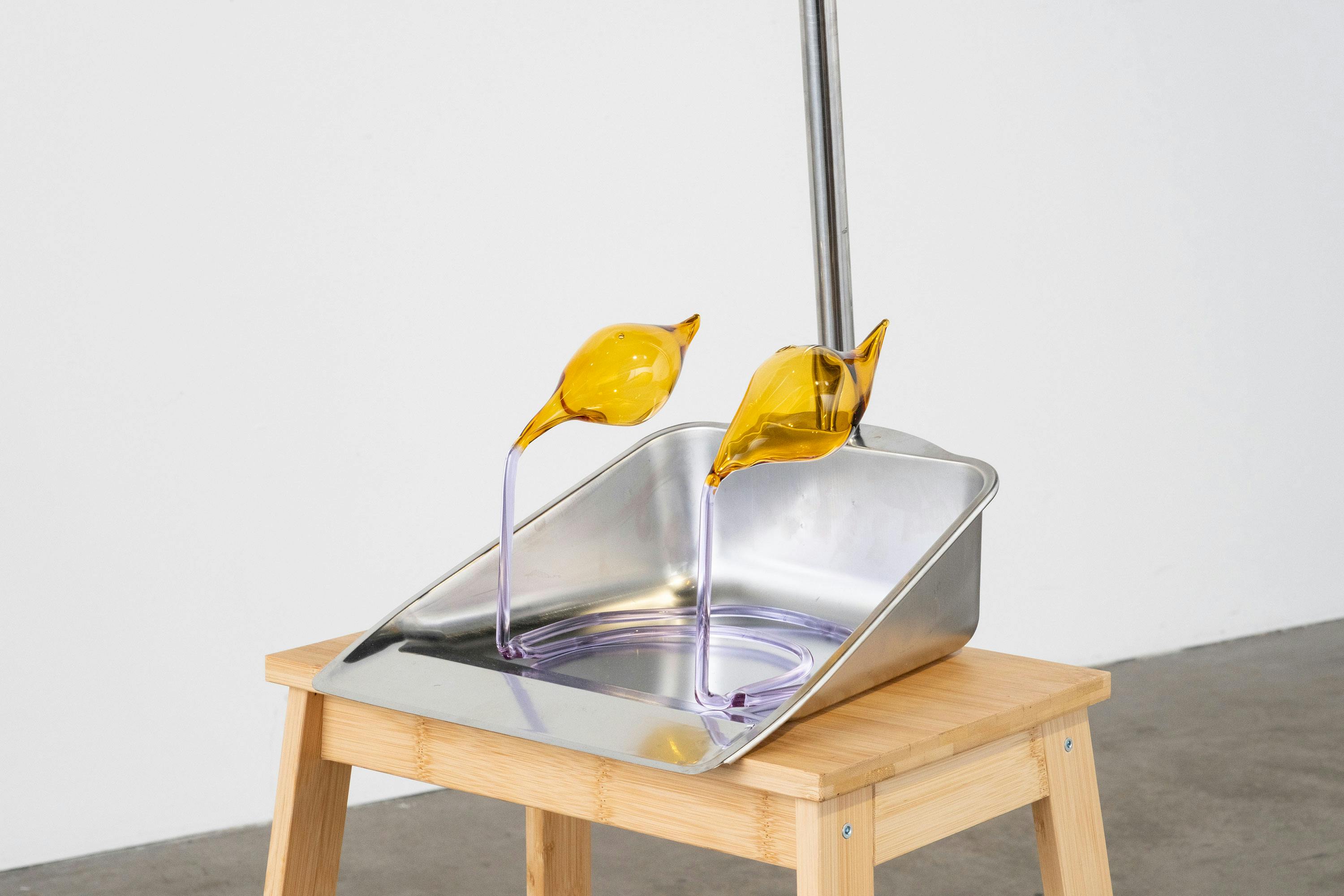 A metal dustpan sits on a wooden stool. Inside the dustpan are two yellow glass sculptures with long, curved purple necks. The dustpan’s handle is upright, and the background is a plain white wall.