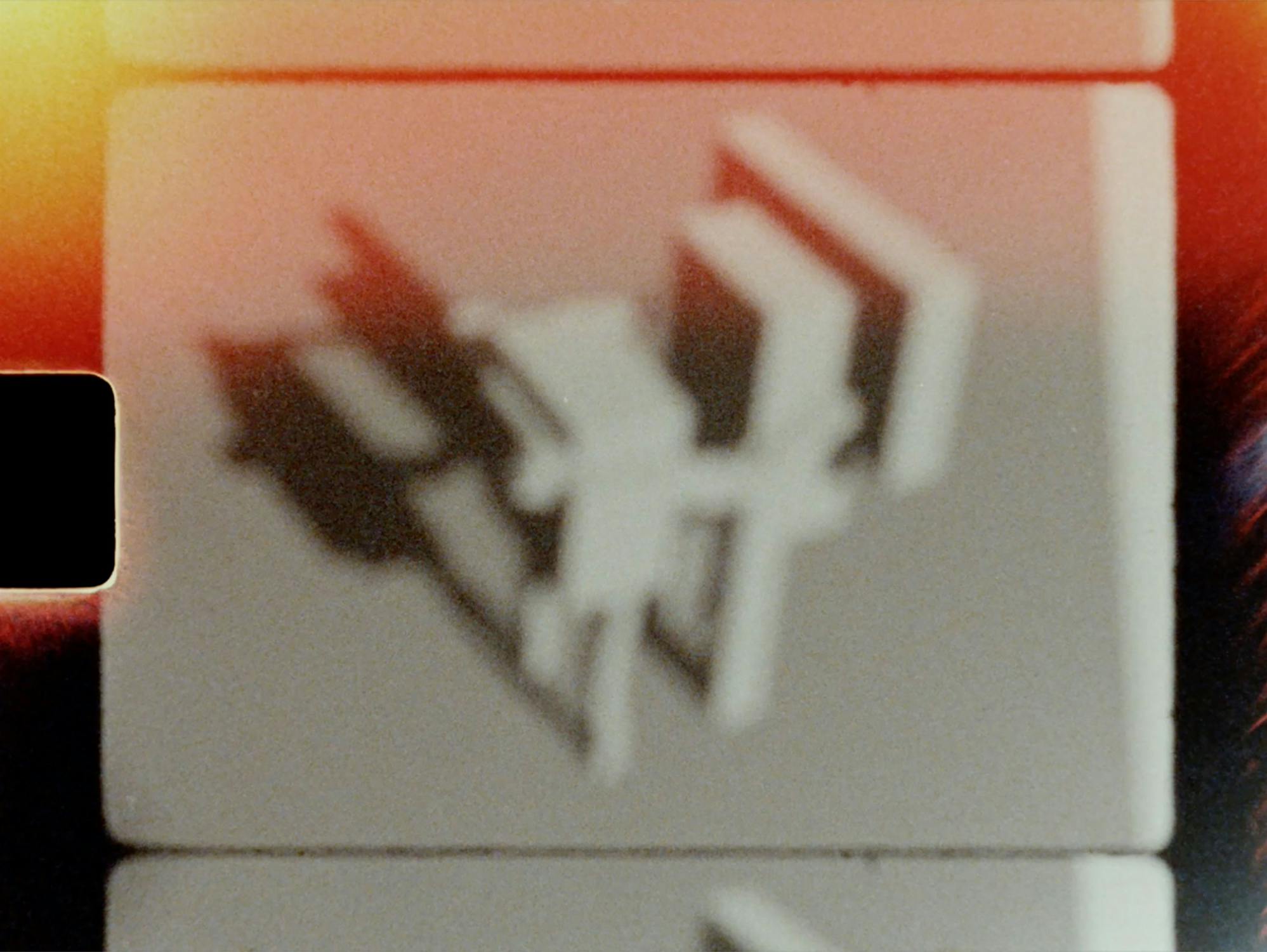A blurred, abstract shadow of a geometric object is cast onto a white surface with a gradient of warm orange light. The scene appears divided by dark horizontal lines, resembling film frames or slides.