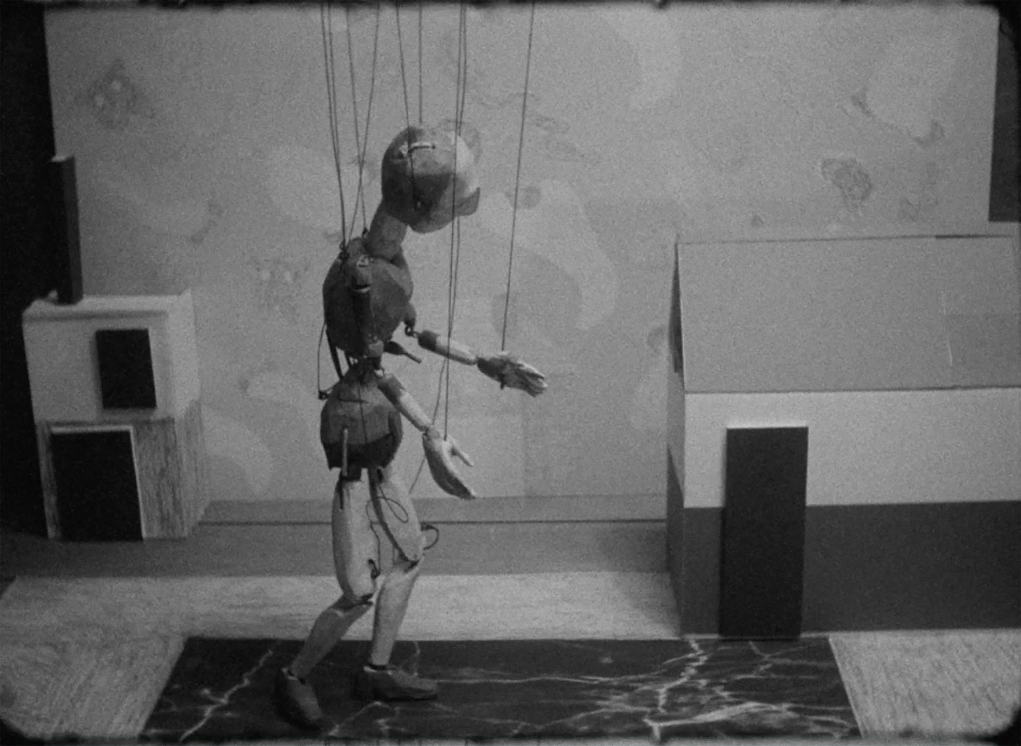 A black-and-white film still shows a wooden marionette puppet walking with outstretched arms. Strings are attached to its limbs and head. The background features boxy, simplistic model buildings and a patterned floor, giving a staged, theatrical appearance.