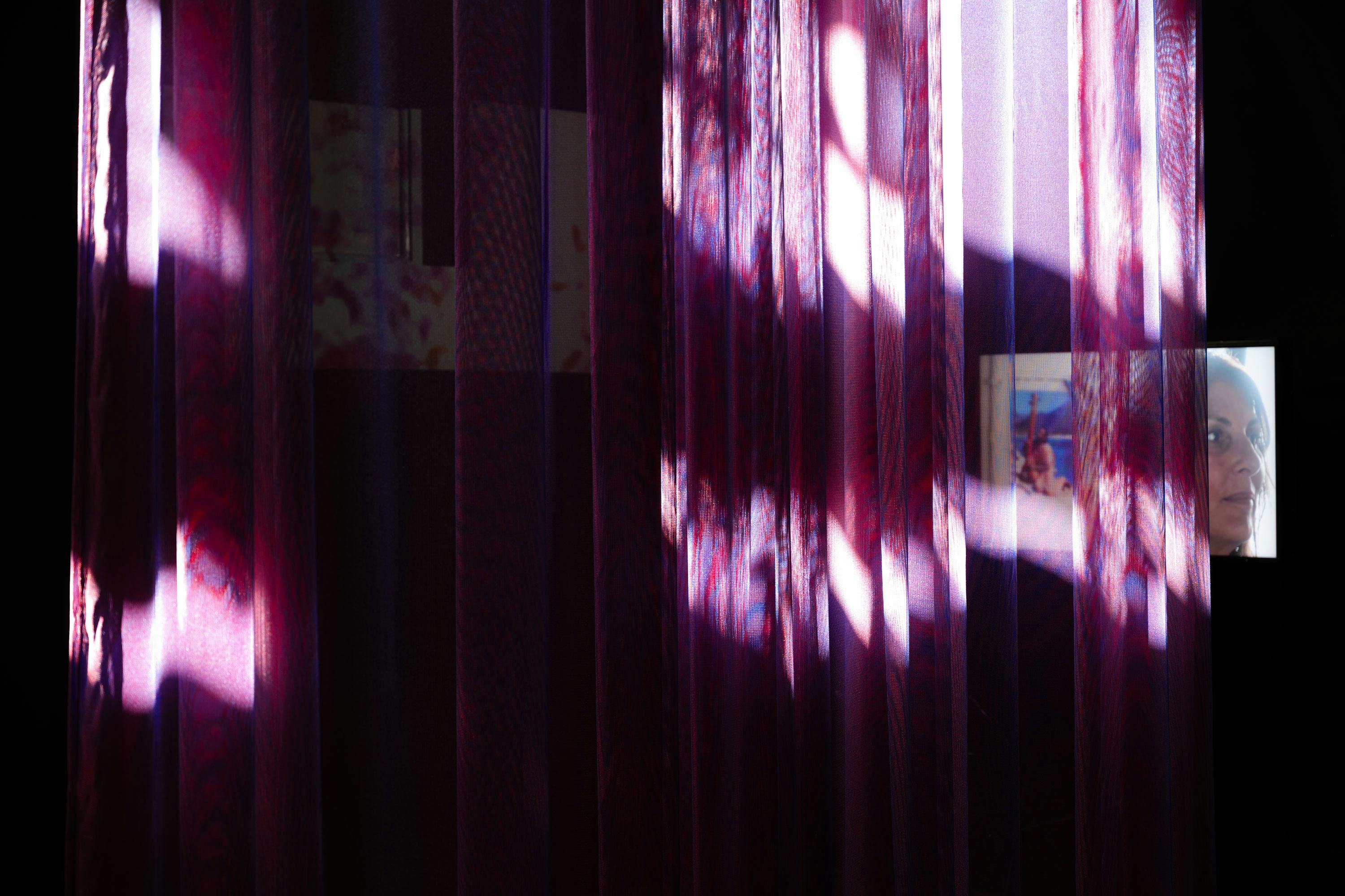 Sunlight streams through sheer, purple curtains with leaf patterns, casting bright patches on the fabric. Behind the curtains, a wall displays blurred images or artworks. In the right background, part of a woman's face is visible in a photo.