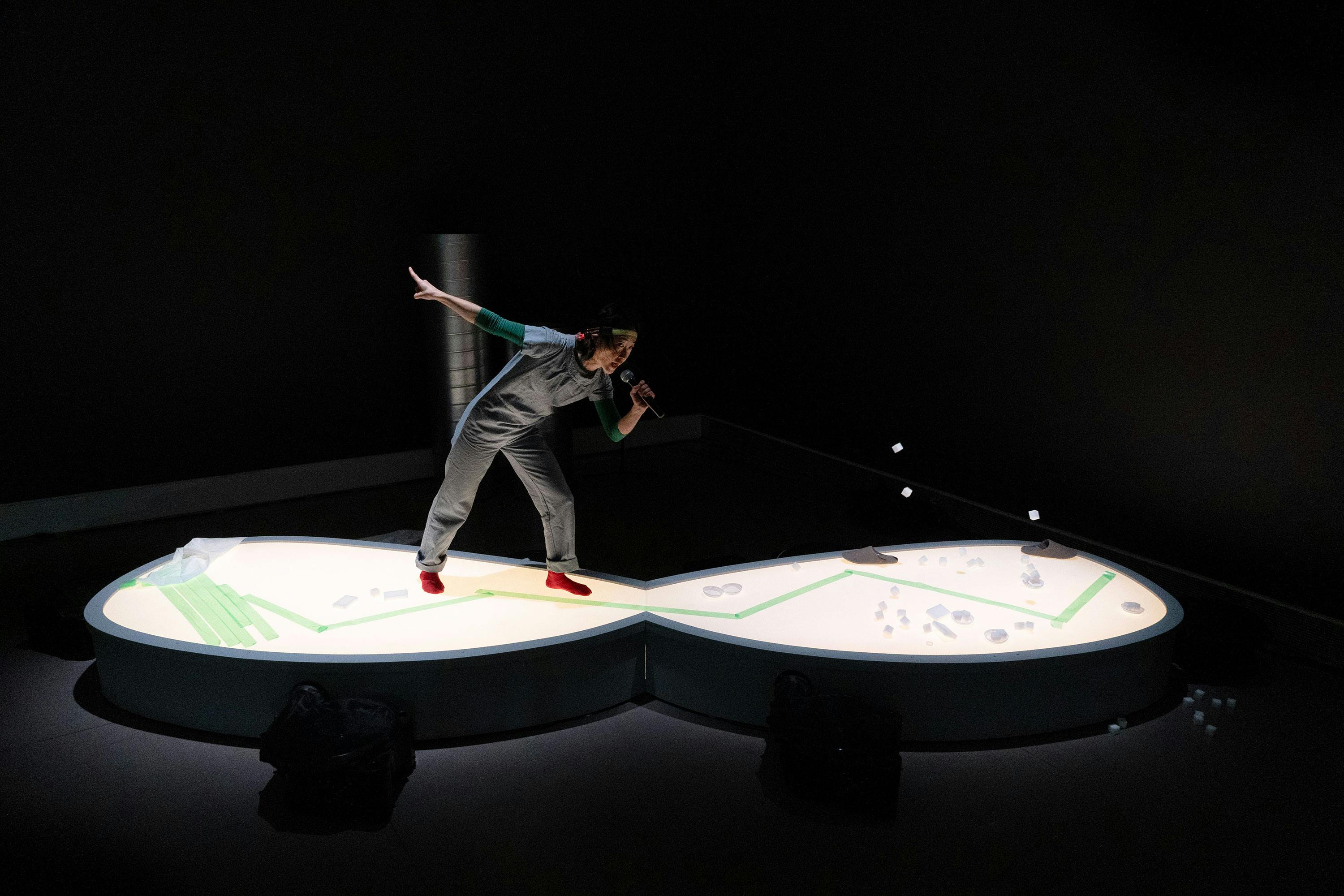 Aki Sasamoto dressed in grey clothing and red shoes standing on a lit, figure-of-eight shaped platform, holding a microphone and gesturing with one arm outstretched, set against a dark background.