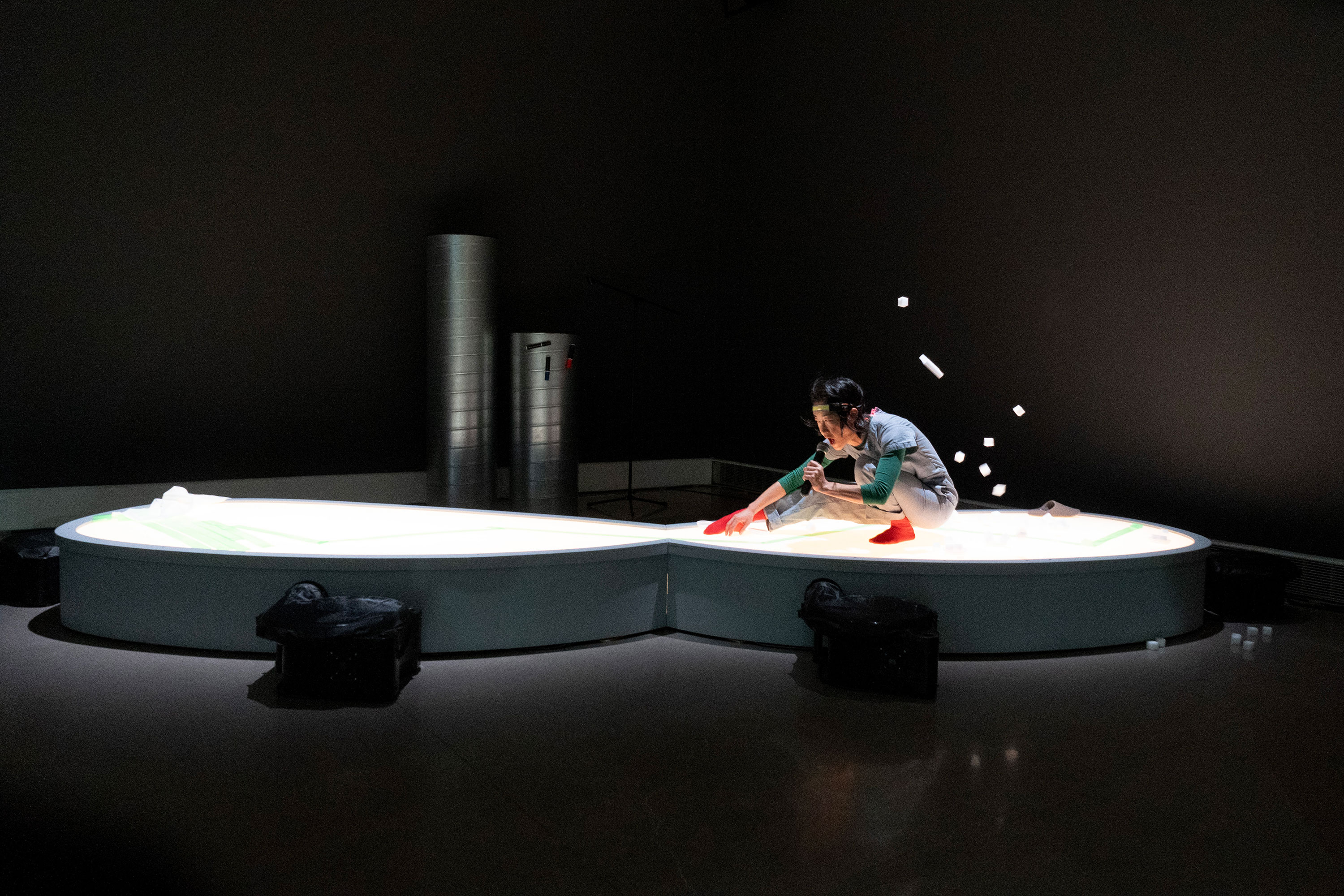 A person kneels on a large, glowing, kidney-shaped platform, arranging objects and tossing white cylindrical items in the air, with scattered objects mid-flight. The dim room features industrial pipes in the background and black bags near the platform’s base.