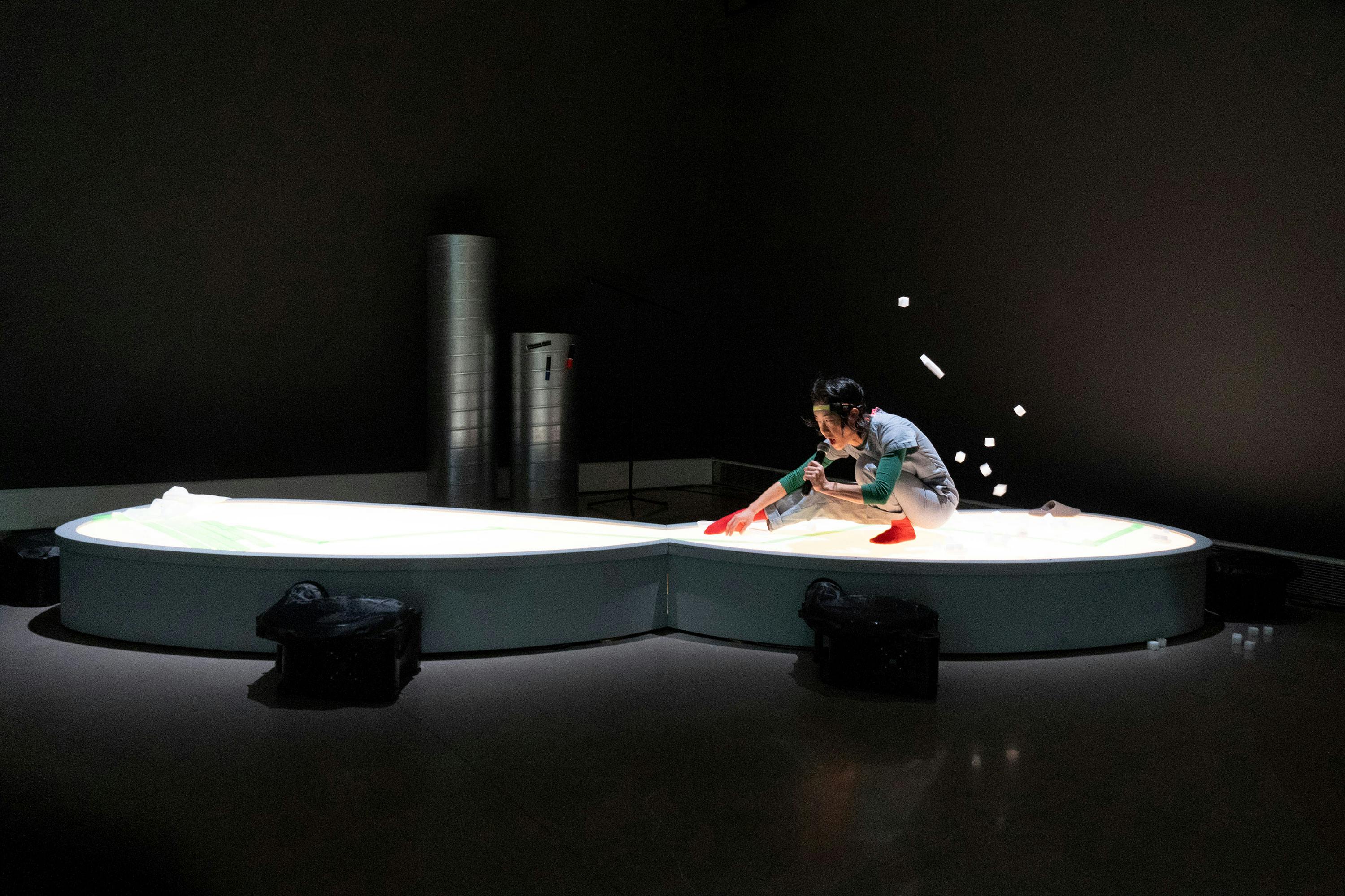 A person kneels on a large, glowing, kidney-shaped platform, arranging objects and tossing white cylindrical items in the air, with scattered objects mid-flight. The dim room features industrial pipes in the background and black bags near the platform’s base.