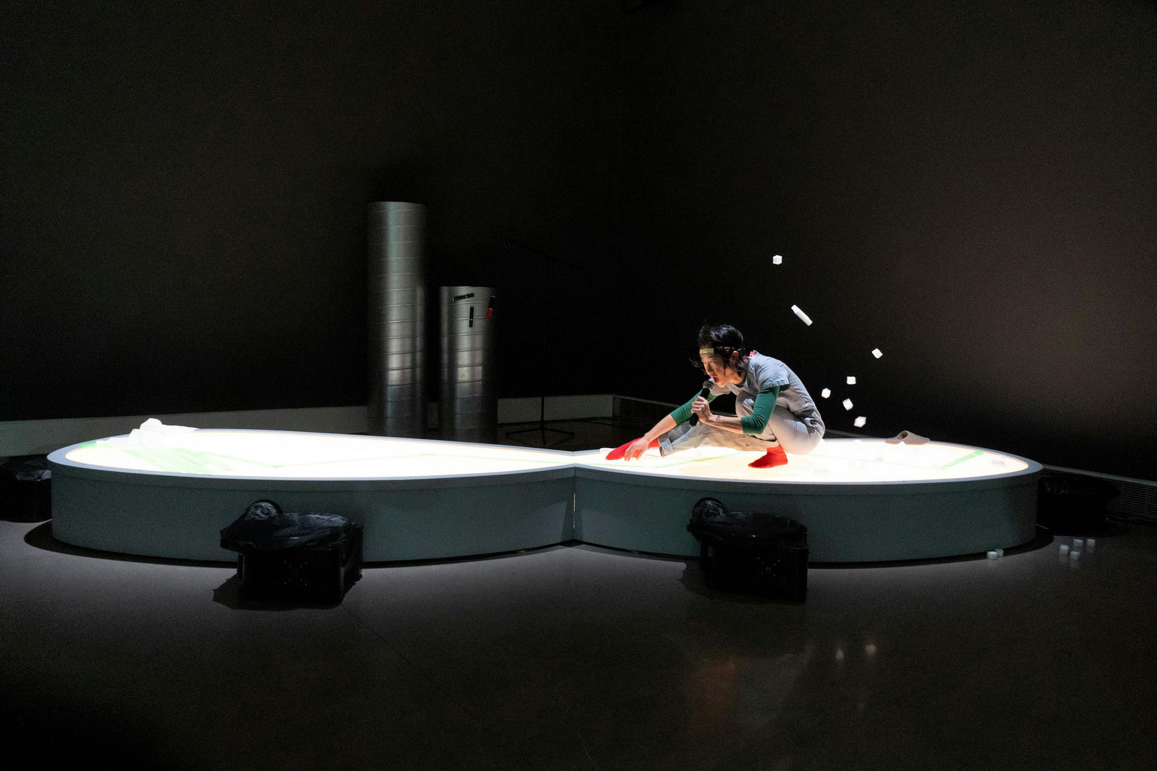 A person kneels on a large, glowing, kidney-shaped platform, arranging objects and tossing white cylindrical items in the air, with scattered objects mid-flight. The dim room features industrial pipes in the background and black bags near the platform’s base.