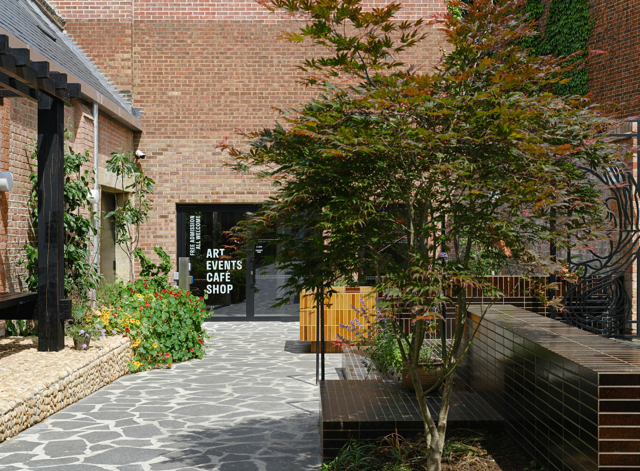 A sunny courtyard with stone paving, wooden benches, green plants, and a small tree. A black door with white lettering stands at the back, and a brick wall forms most of the background. The garden looks inviting and well-maintained.