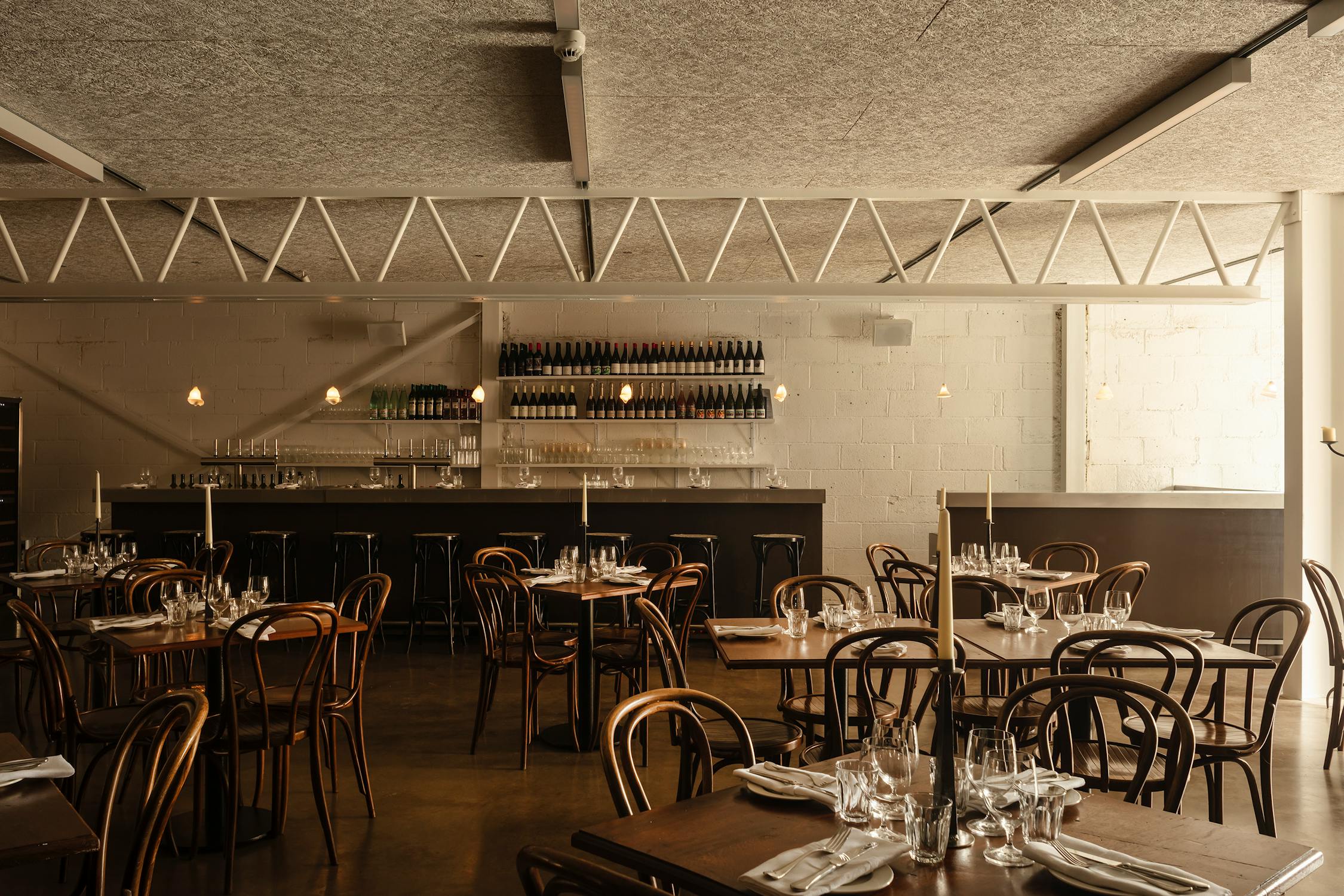 A warmly lit, modern restaurant with wooden tables and chairs arranged neatly. Tables are set with glasses, plates, and cutlery. In the background, there’s a bar with shelves holding bottles and glasses, and minimalist pendant lights hang above.