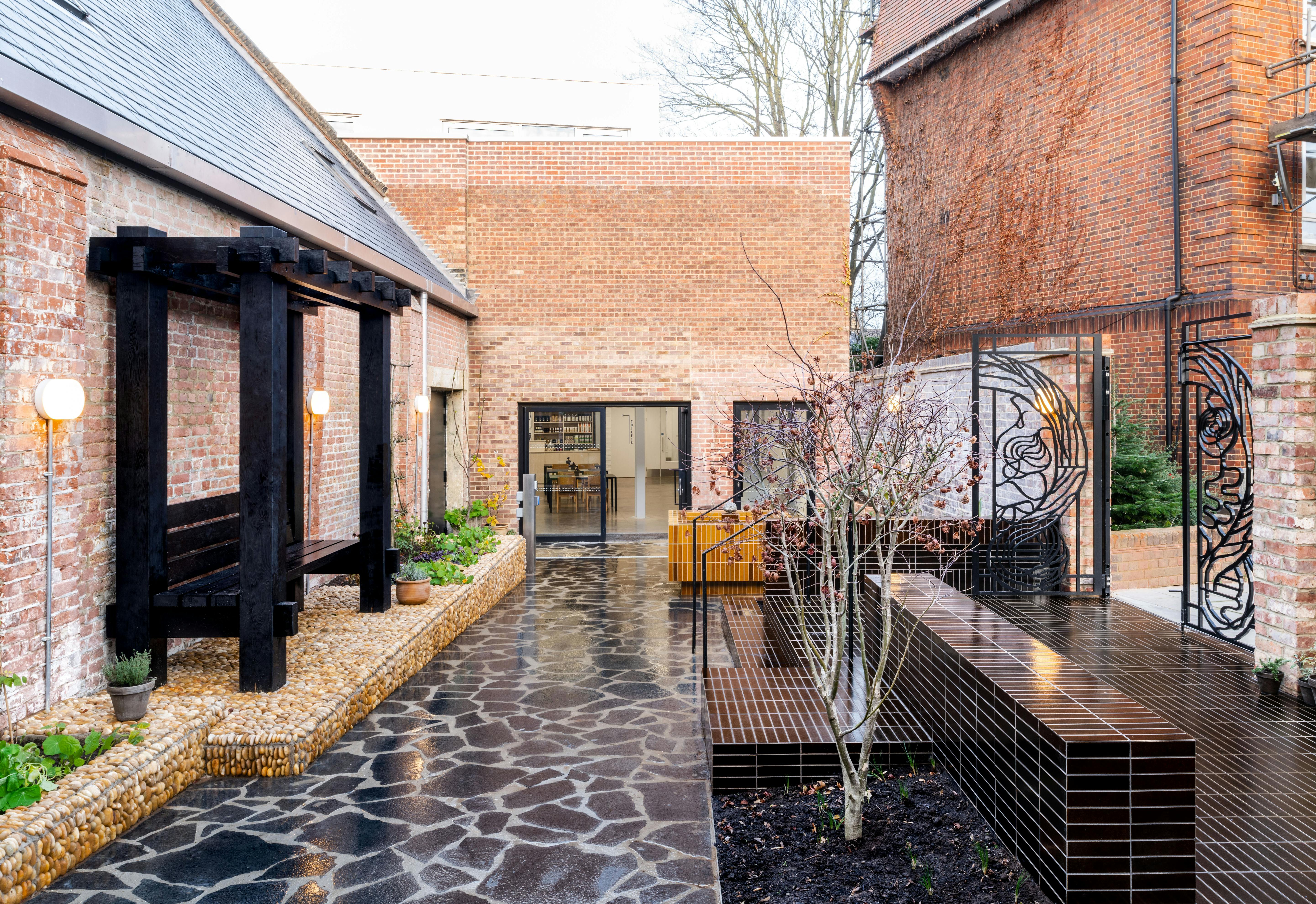 A modern courtyard with brick walls, black stone path, raised planters, bare trees, decorative metal panels, and globe wall lights. The space blends rustic brick with sleek, contemporary design elements under soft, natural daylight.