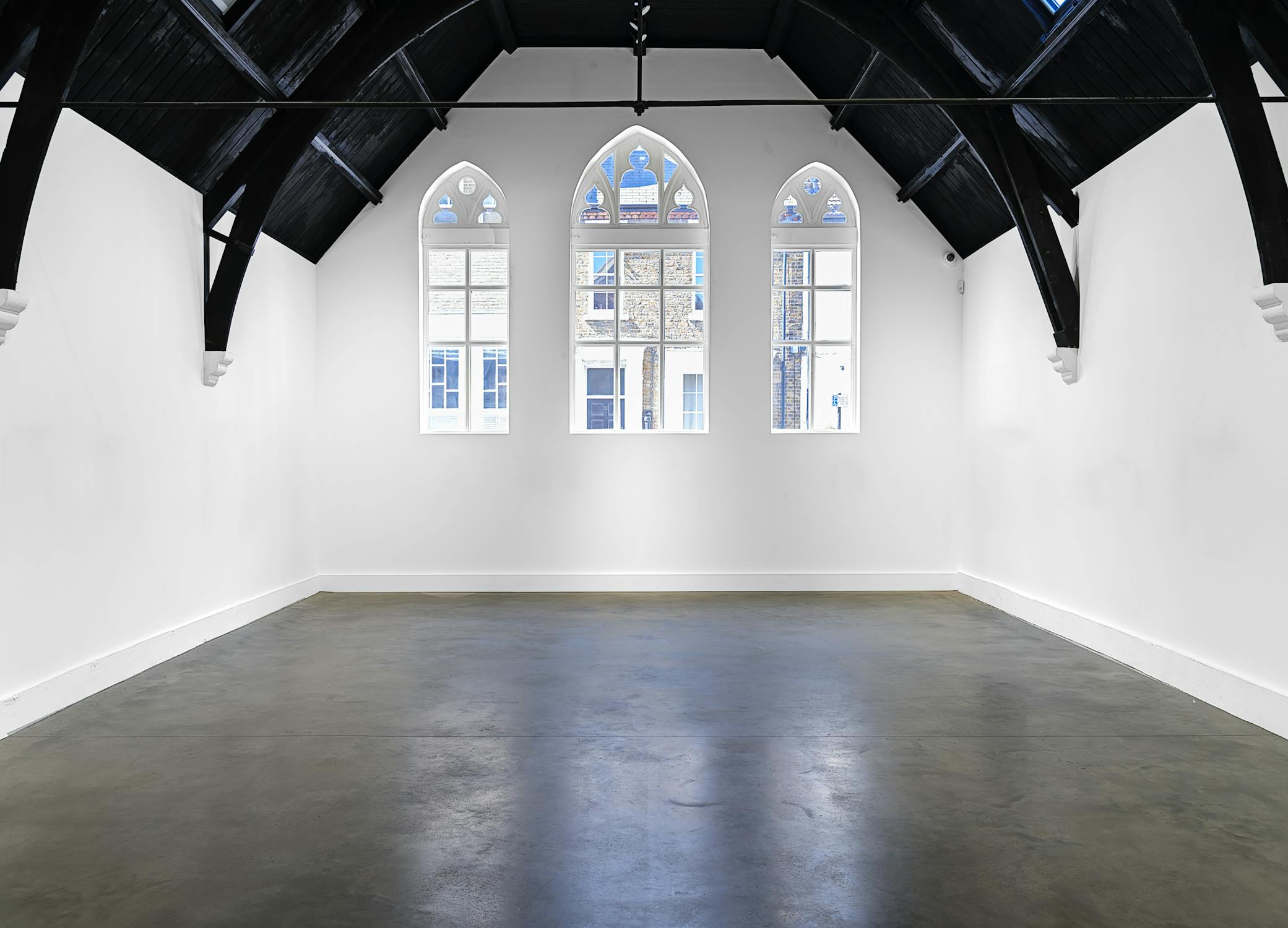 A bright, empty gallery room with white walls, a polished concrete floor, and a vaulted black ceiling. Three tall, arched windows line the far wall, letting in natural light and revealing a building outside. The space feels open and modern.