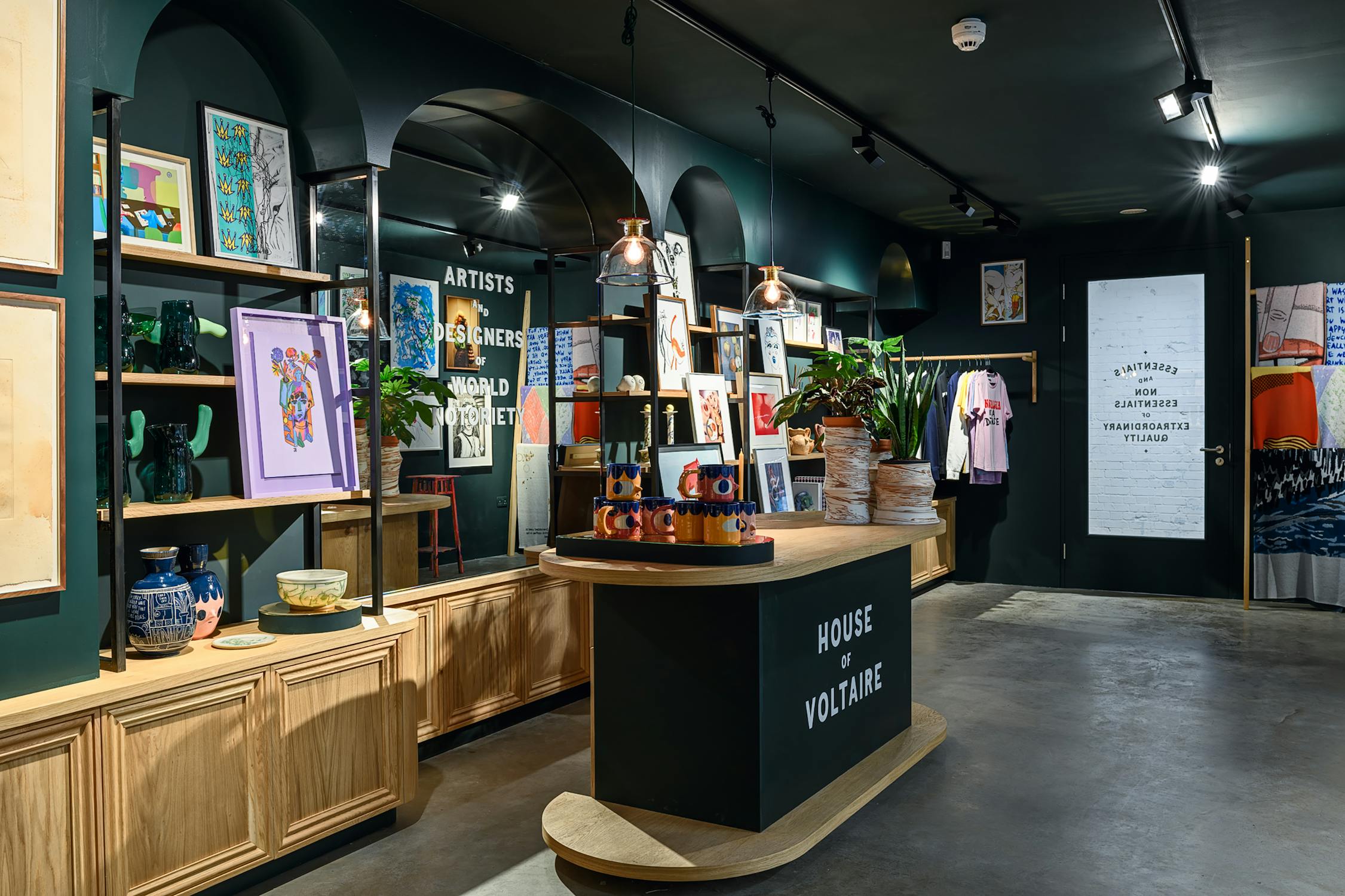 A modern shop features framed colourful artwork on dark green walls and wooden shelves. A black reception desk with “House of Voltaire” signage stands in the foreground. Spotlights and potted plants add warmth to the stylish, contemporary space.