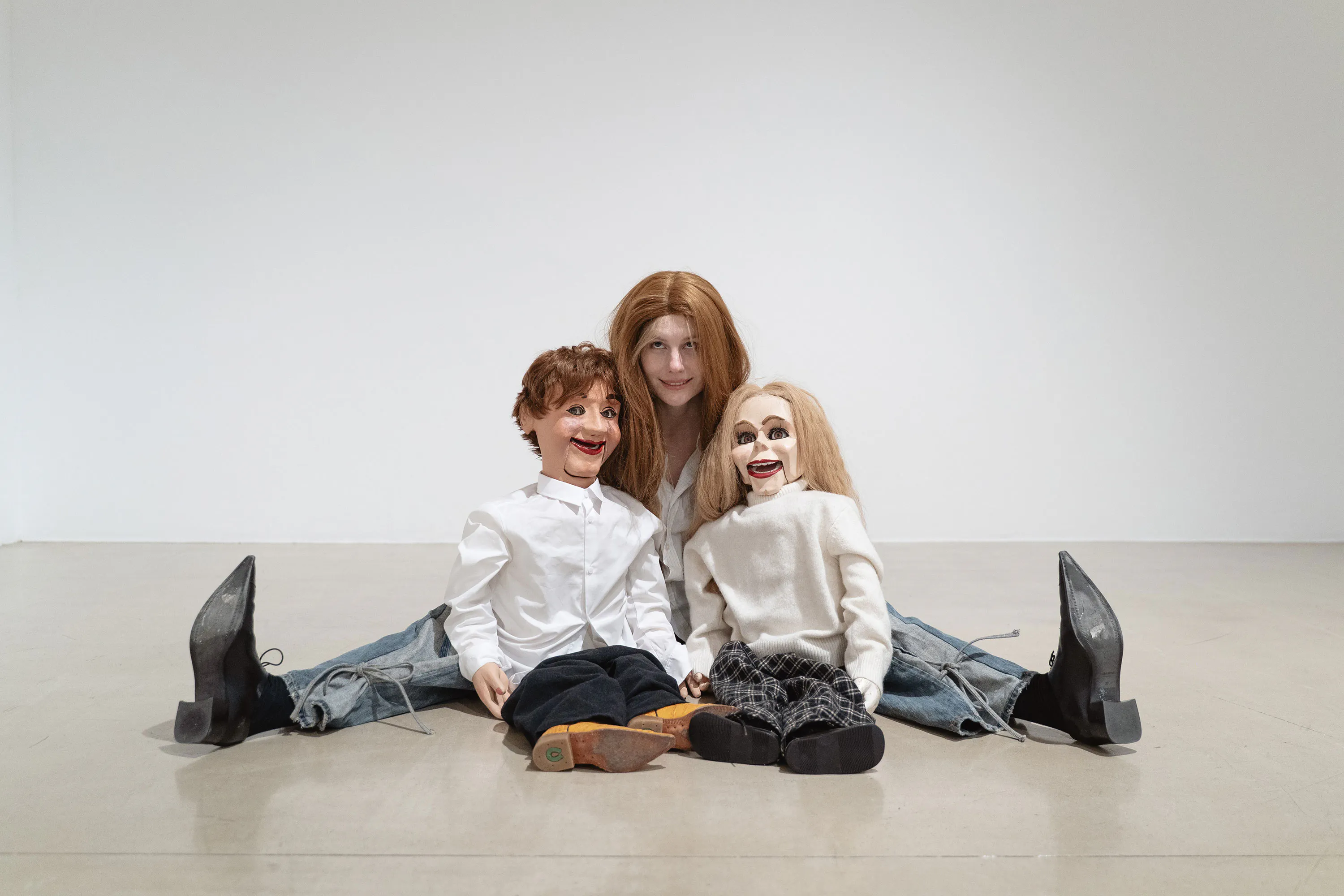 Stina Fors sits on the floor against a white wall, legs spread wide. In front of them are two ventriloquist dummies with exaggerated smiles, dressed in white tops and dark trousers, positioned to mimic the person’s seated posture.