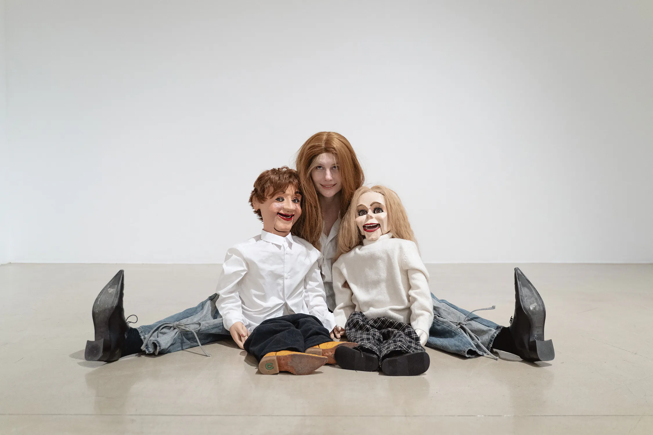 Stina Fors sits on the floor against a white wall, legs spread wide. In front of them are two ventriloquist dummies with exaggerated smiles, dressed in white tops and dark trousers, positioned to mimic the person’s seated posture.