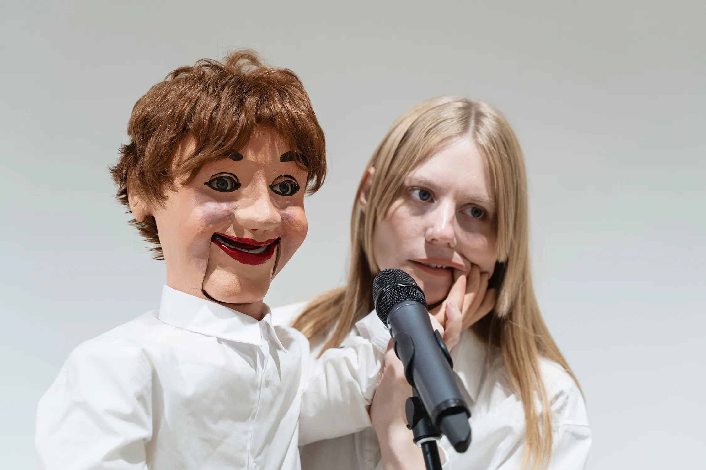 Stina Fors stands behind a ventriloquist dummy with brown hair and exaggerated facial features, both facing forwards. The puppet's hand touches the person’s cheek, and they both stand near a black microphone against a plain background.