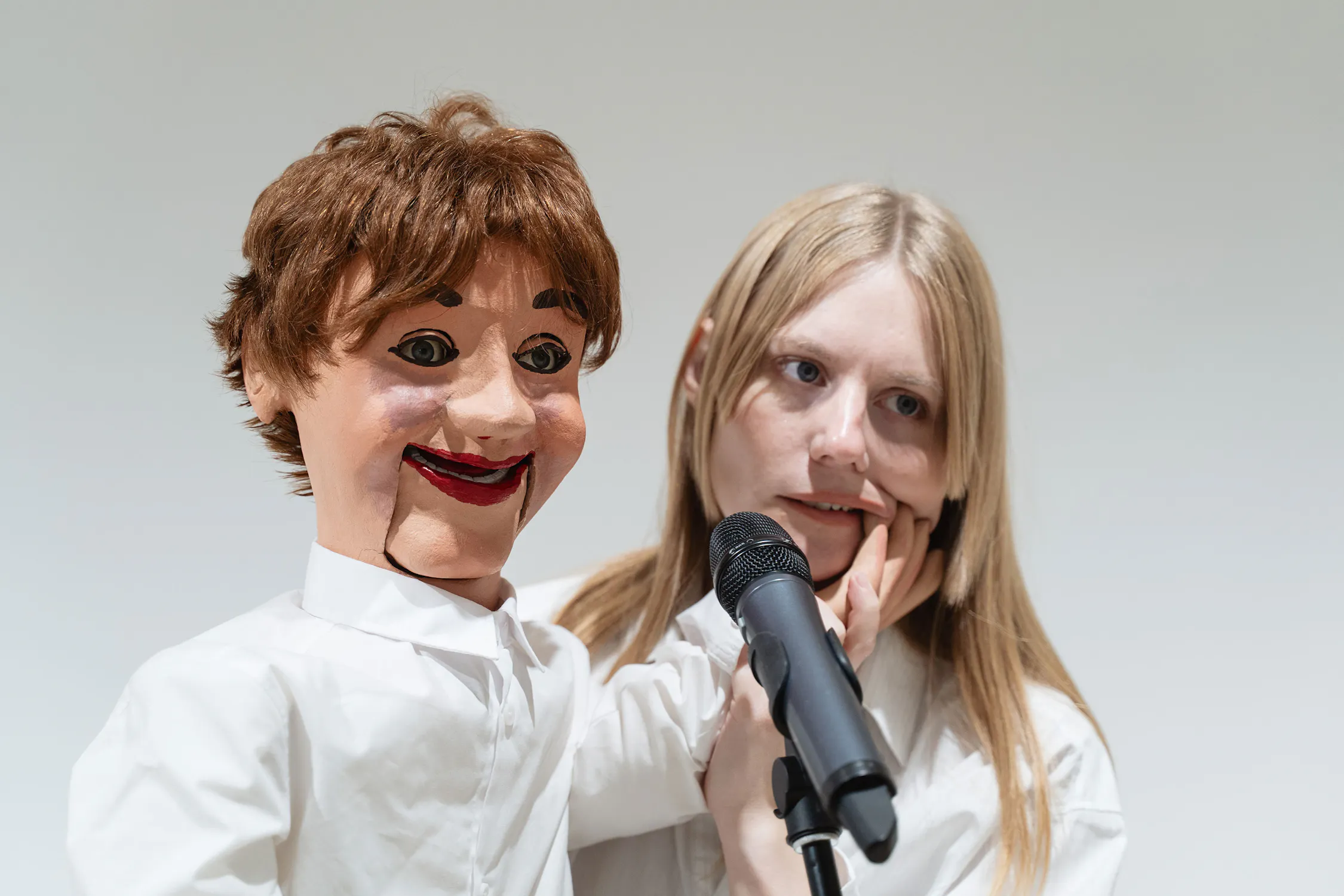 Stina Fors stands behind a ventriloquist dummy with brown hair and exaggerated facial features, both facing forwards. The puppet's hand touches the person’s cheek, and they both stand near a black microphone against a plain background.