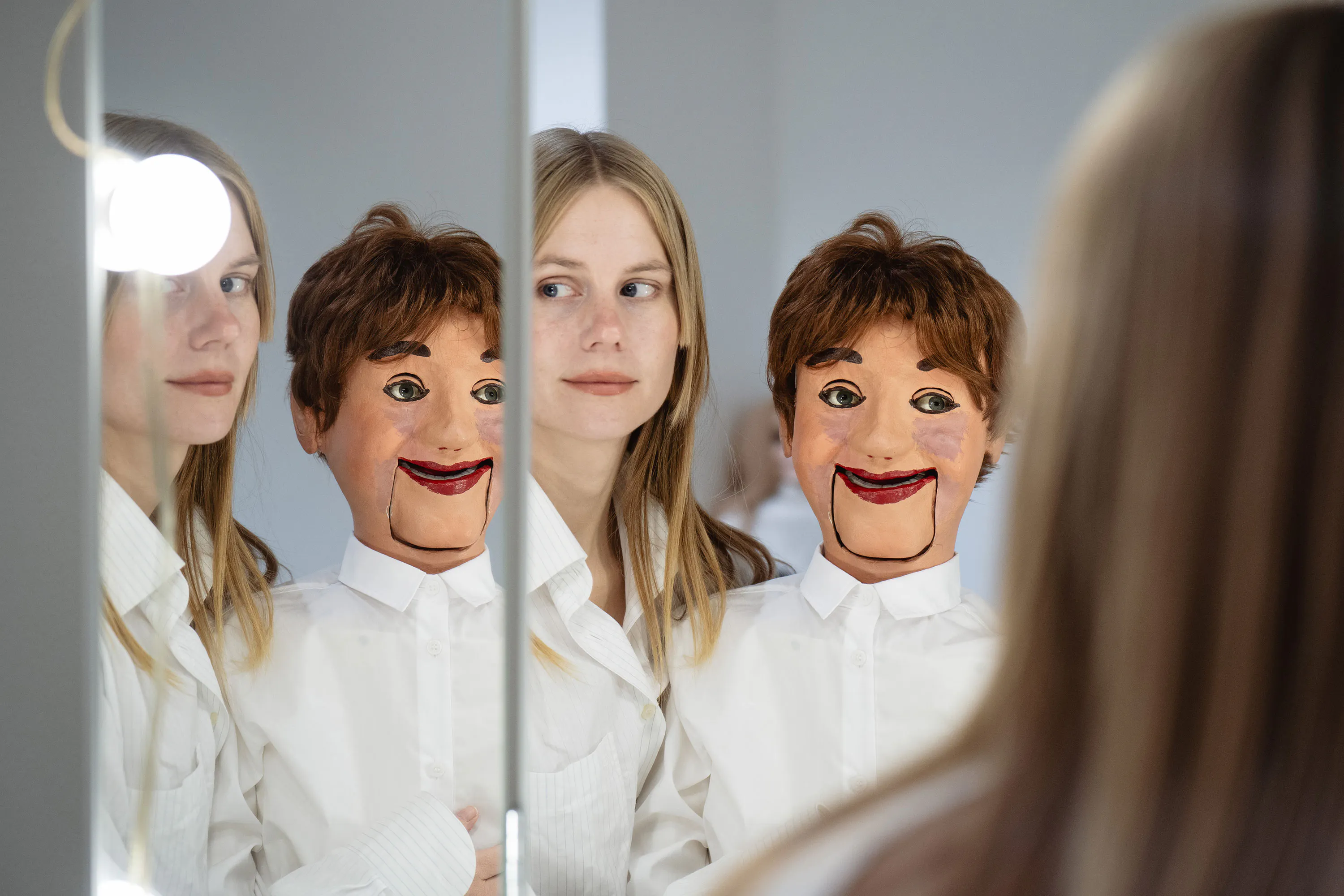Stina Fors, wearing a white shirt, stands next to a ventriloquist's dummy with brown hair and a wide smile. Both are reflected in a mirror, creating multiple images of them in the scene. The background is softly lit and minimalist.