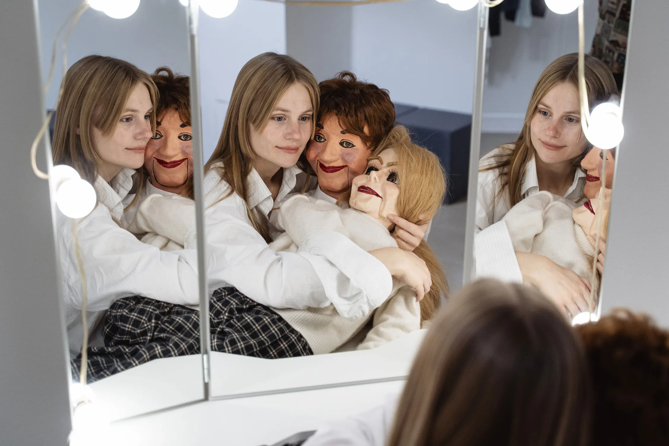 A woman with light brown hair sits at a dressing table mirror, hugging two ventriloquist dummies. She smiles softly, and her reflection is visible in three mirror panels. The dummies have painted faces and wigs, dressed in light shirts. Round lights frame the mirror.