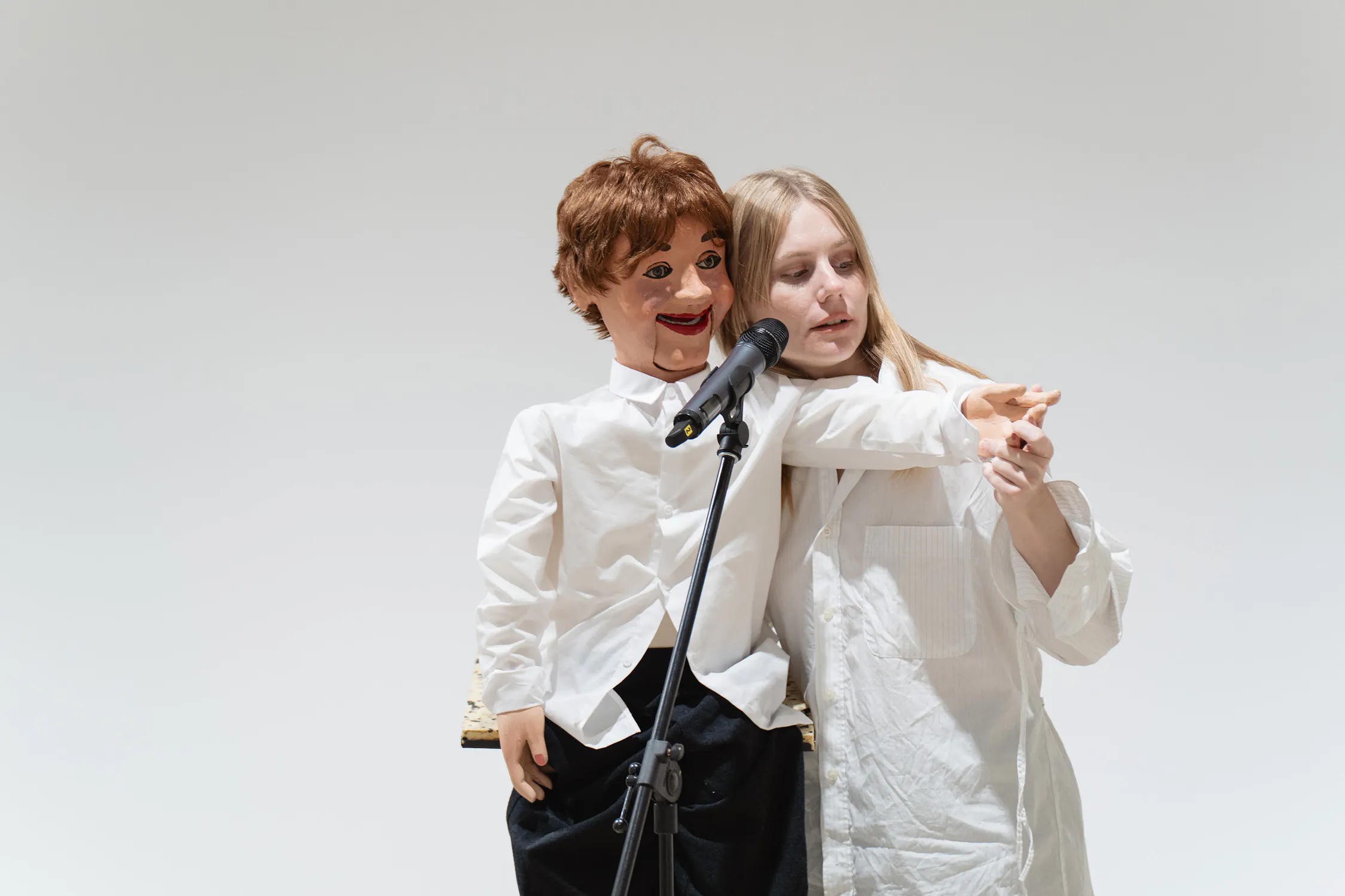 Artist Stina Fors operates a brown-haired ventriloquist dummy, also dressed in white, at a microphone stand. The person’s arm gestures forward, guiding the puppet’s hand to point as they perform together against a plain white background.