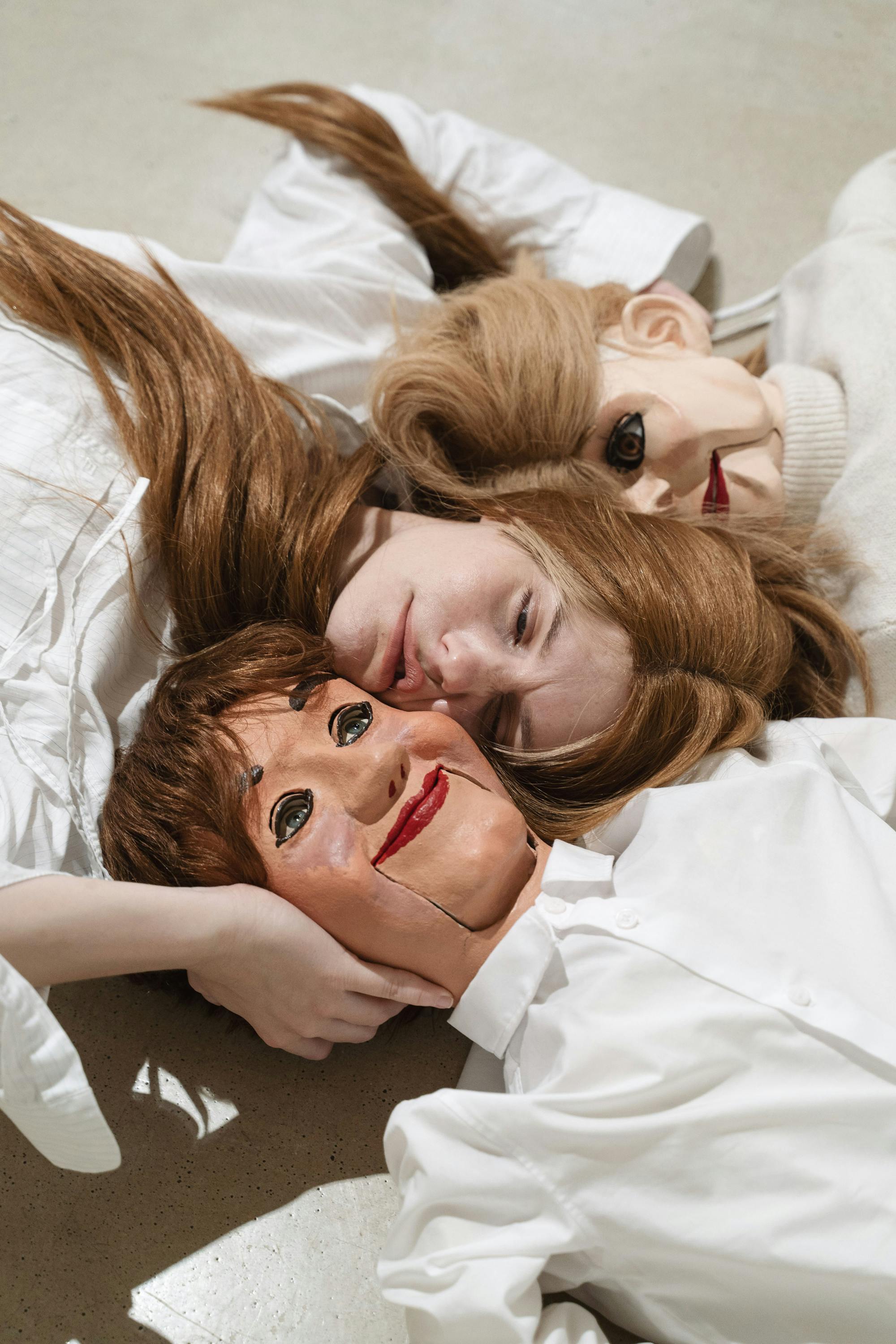 Artist Stina Fors lies on the floor, embracing two ventriloquist dummies or masks with painted faces and synthetic hair. The person is dressed in white, blending into the neutral background, and gazes softly at the camera.