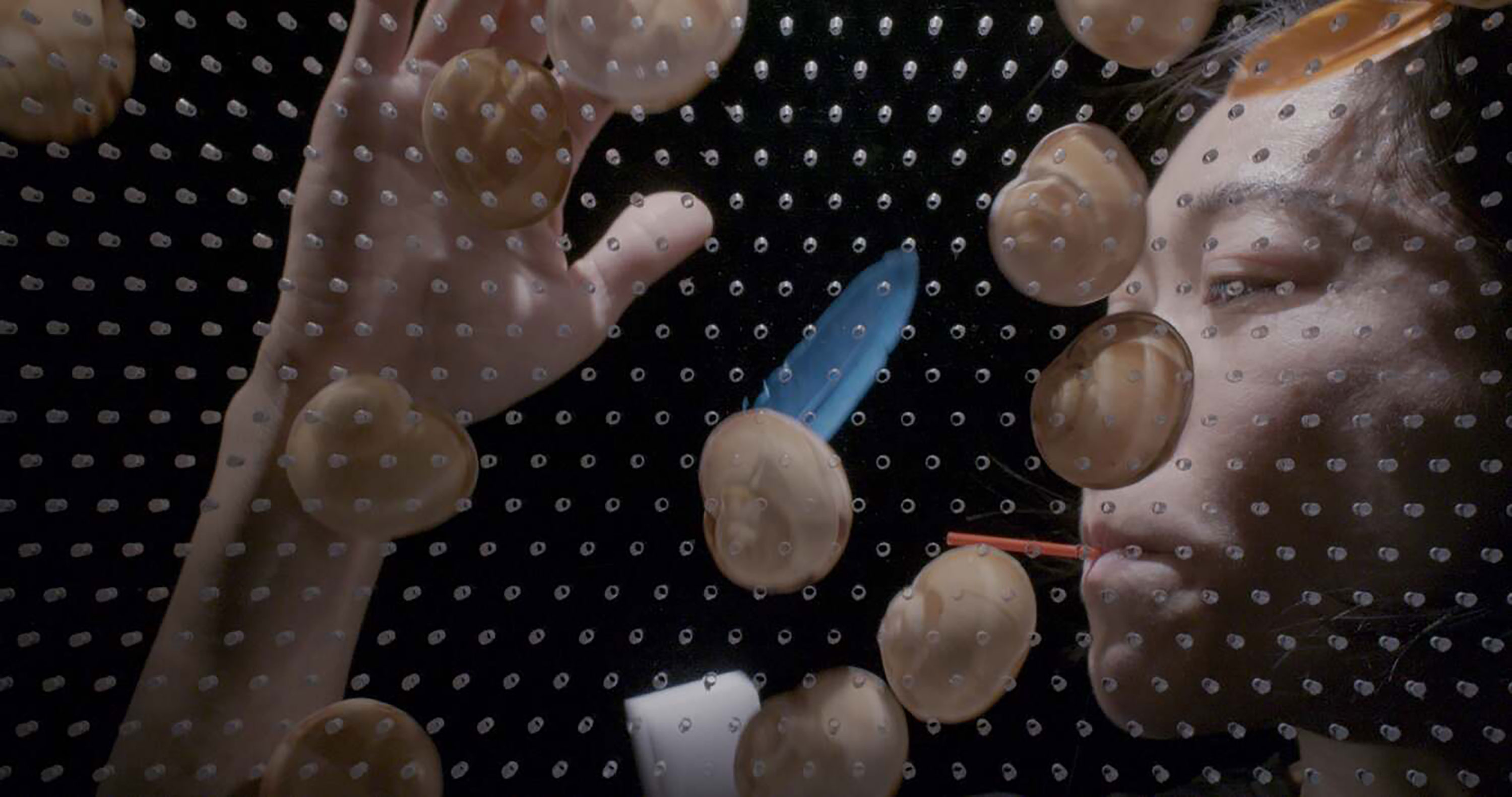 A person presses their face and hand against a glass panel with many small suction pads. Their eyes are closed, and their mouth is distorted by a red object. The background is dark, making the face, hand, and suction pads stand out dramatically.