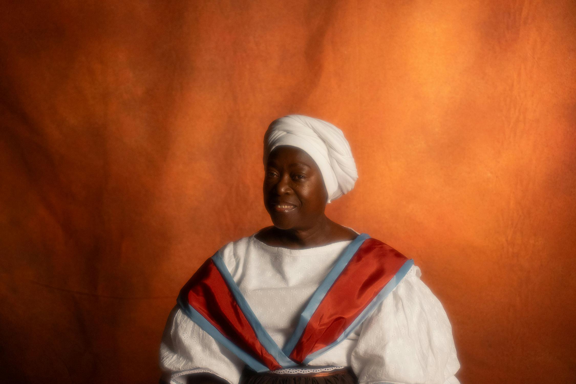 An elderly Black woman in traditional dress sits between two candlesticks against a warm, brown backdrop. She wears a white headscarf, a white blouse with red and blue shawl, and a long dark skirt. She looks slightly to the side with a gentle expression.