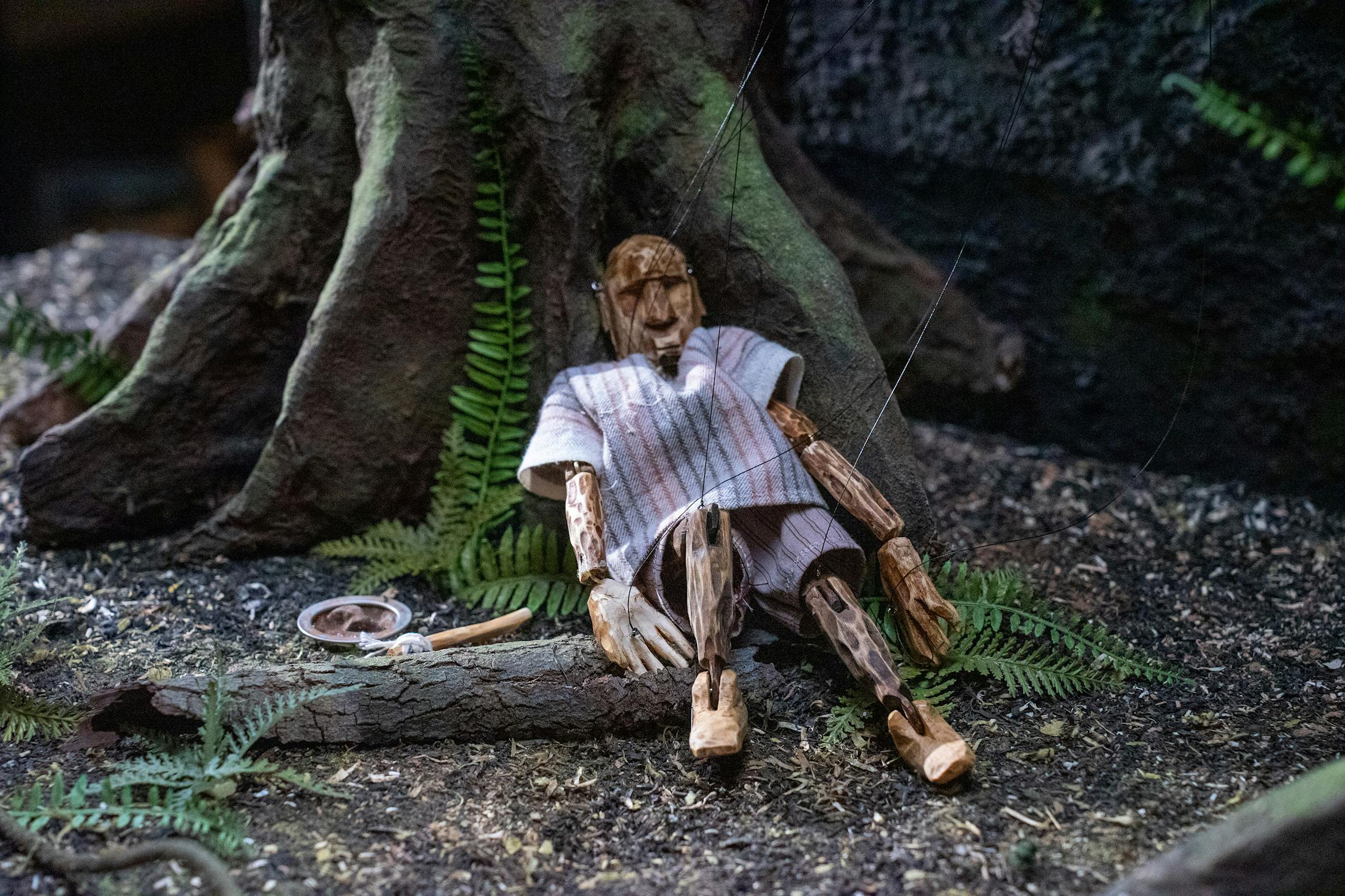 A wooden marionette wearing a striped tunic sits slumped against a large tree root, surrounded by ferns and earthy ground. One of its strings appears cut, with the control handle lying nearby, suggesting it is inactive or at rest in a forest setting.