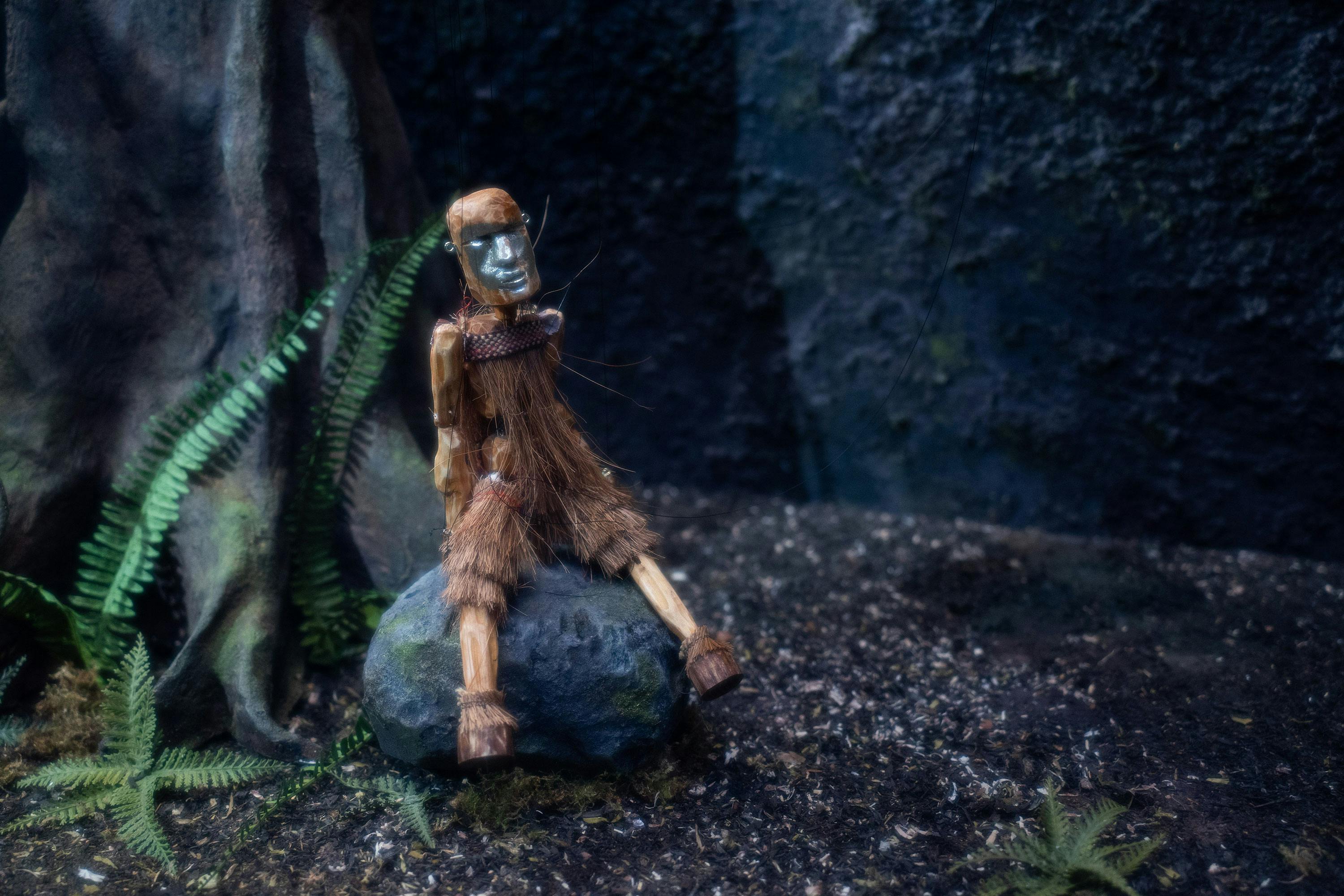 A small wooden figure, dressed in fur-like material and wearing a mask, sits on a rock beside a tree. Green ferns grow nearby on the dark, textured forest floor. The dim, eerie lighting creates a mysterious, woodland atmosphere.