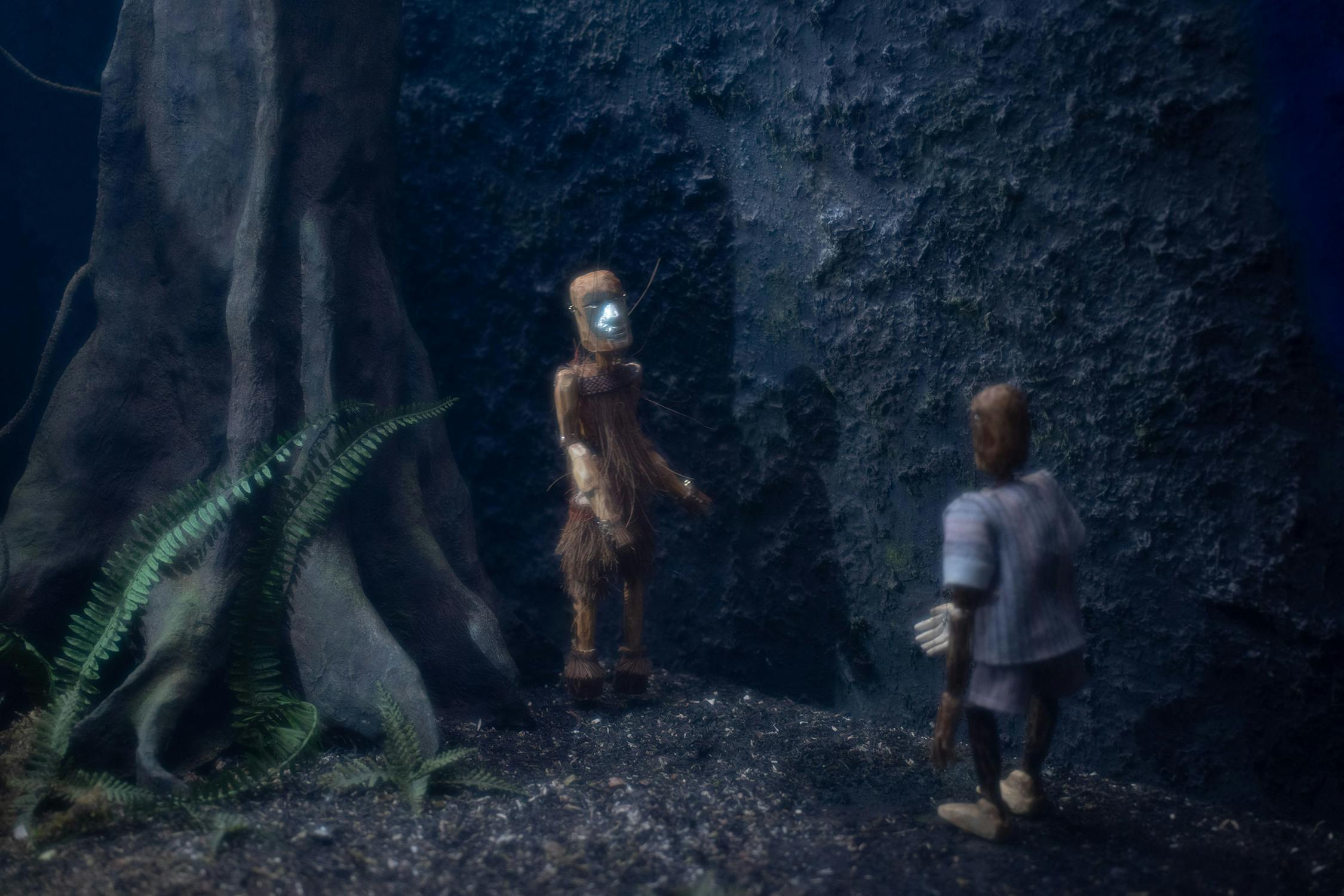 Two small humanoid figurines stand in a dark, forest-like setting. One figure, dressed in brown with a blue face, stands near a large tree and ferns. The other figure, in grey, faces the first against a textured, rocky background. The scene appears dimly lit.