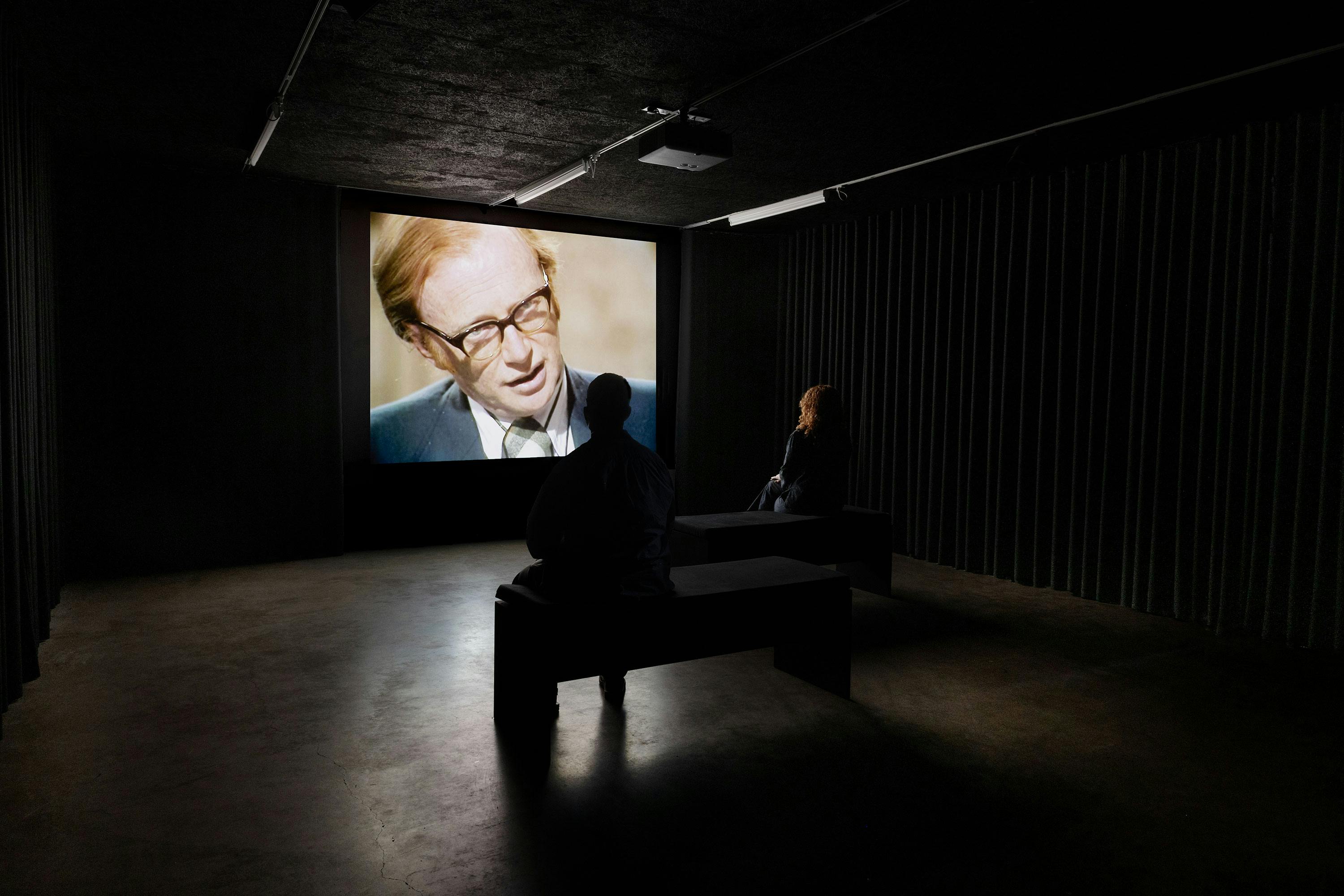 Two people sit on benches in a dark room, watching a film projected on a large screen. The screen shows a close-up of Dennis Potter, a white man in glasses and a suit speaking