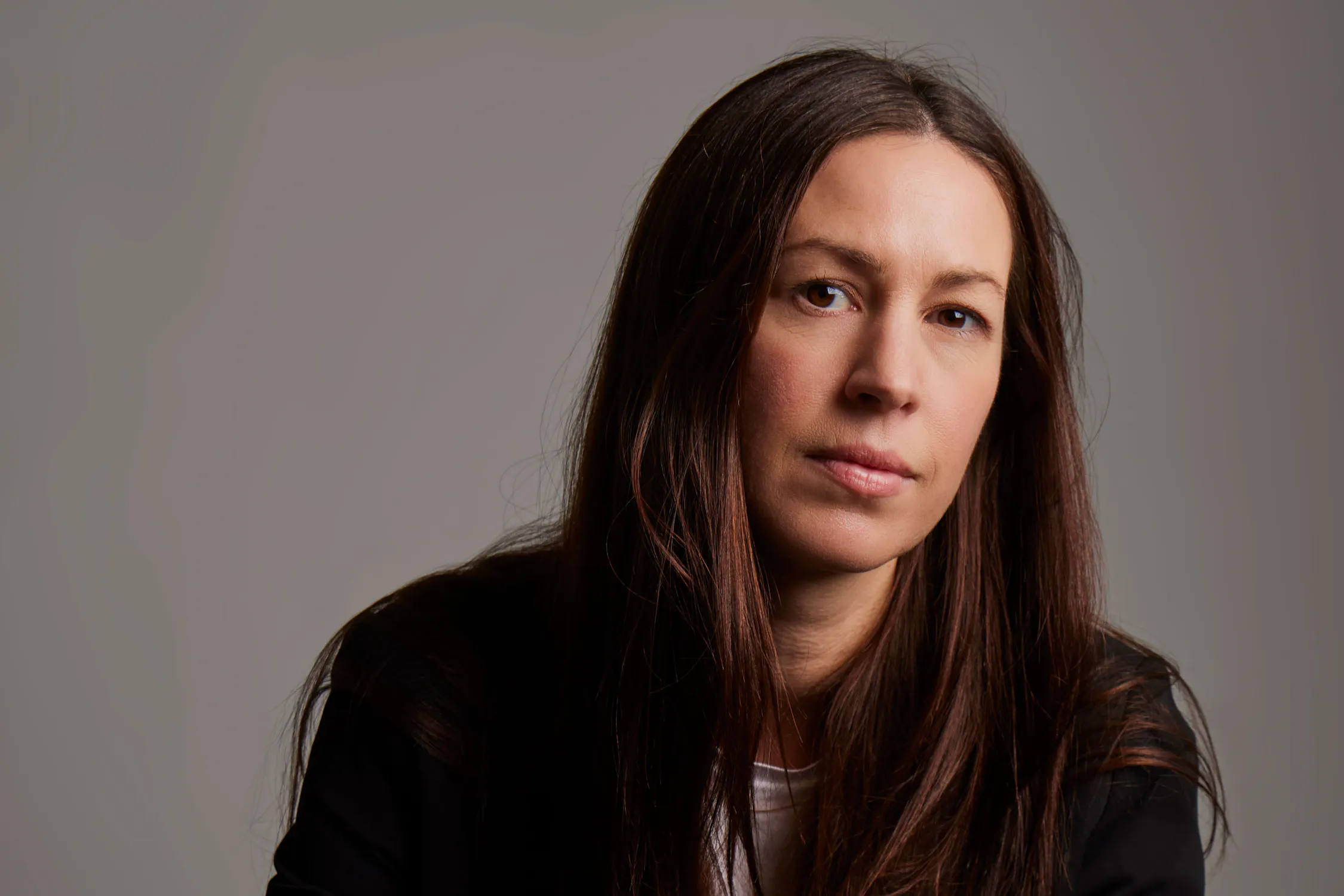 A woman with long brown hair and a neutral expression looks directly at the camera. She wears a white shirt under a black jacket. The background is plain and grey, and the lighting is soft, highlighting her face and hair.