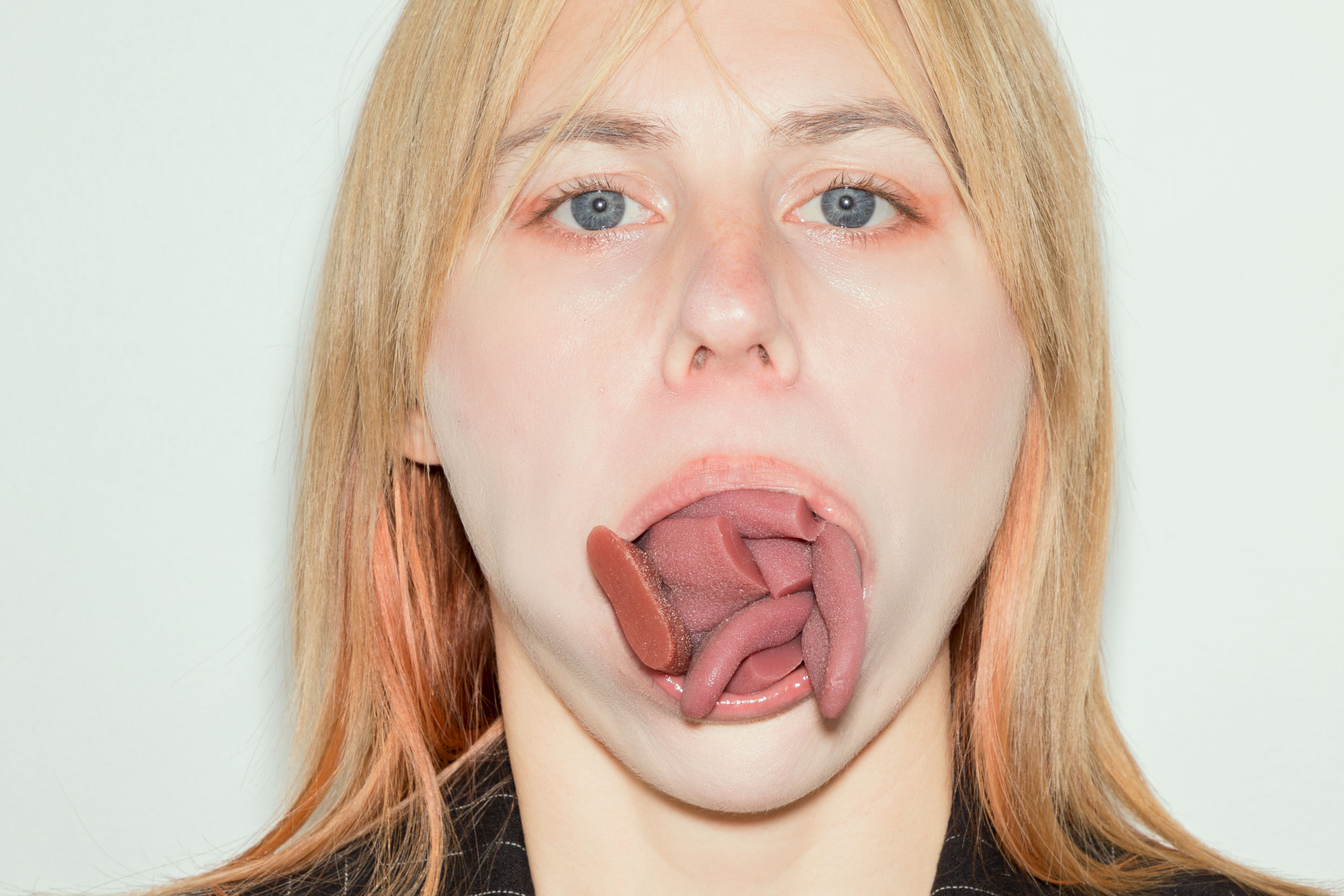 Stina Fors stares blankly at the camera. Their mouth is open, revealing multiple realistic-looking tongue models stacked inside, creating a surreal and unsettling effect. The background is plain and white.