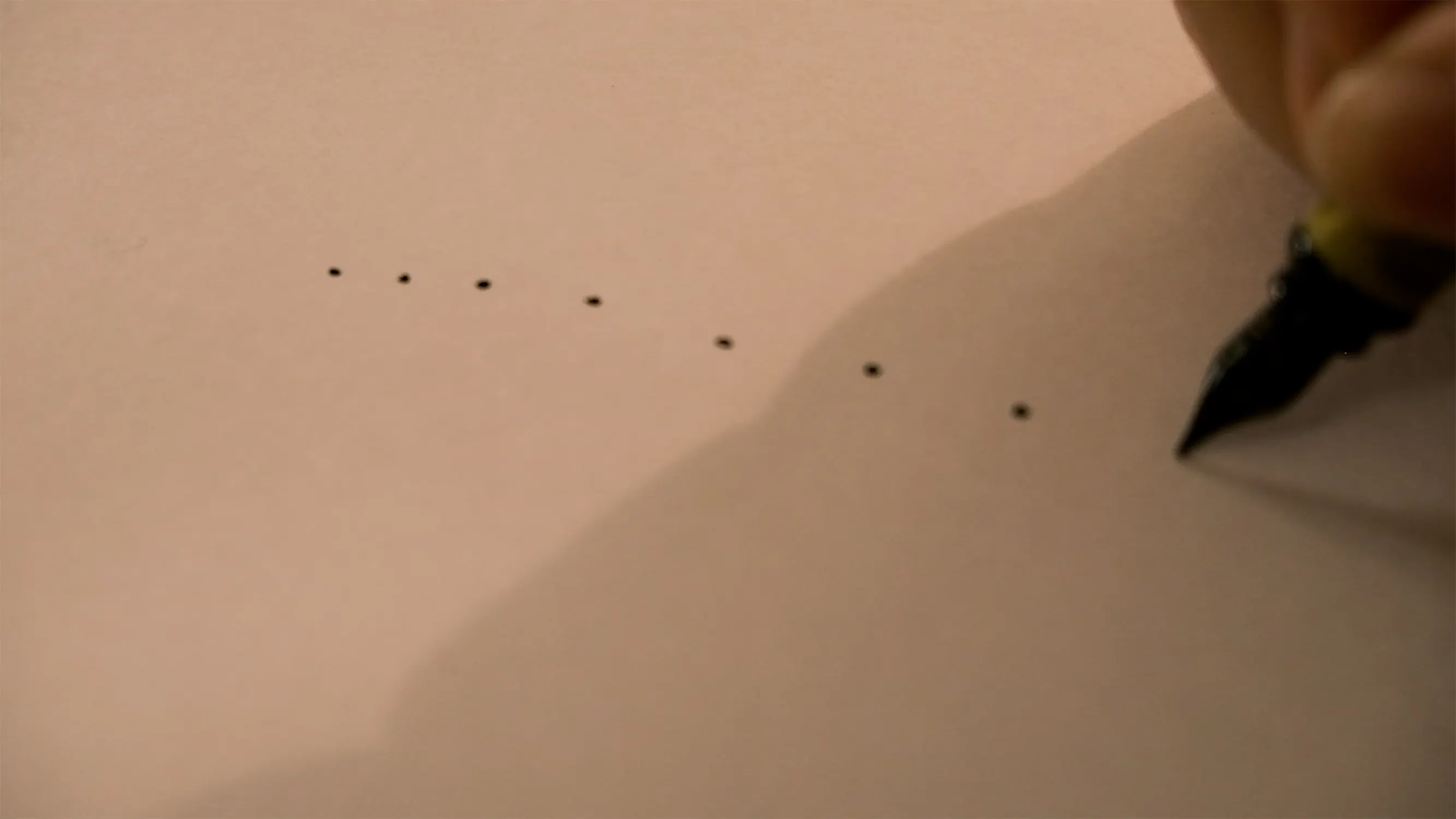 A close-up of a fountain pen held by a hand, drawing a series of small black dots in a line on a plain sheet of paper. The lighting casts a shadow from the hand onto the paper, and the focus is on the pen tip and the dotted pattern.