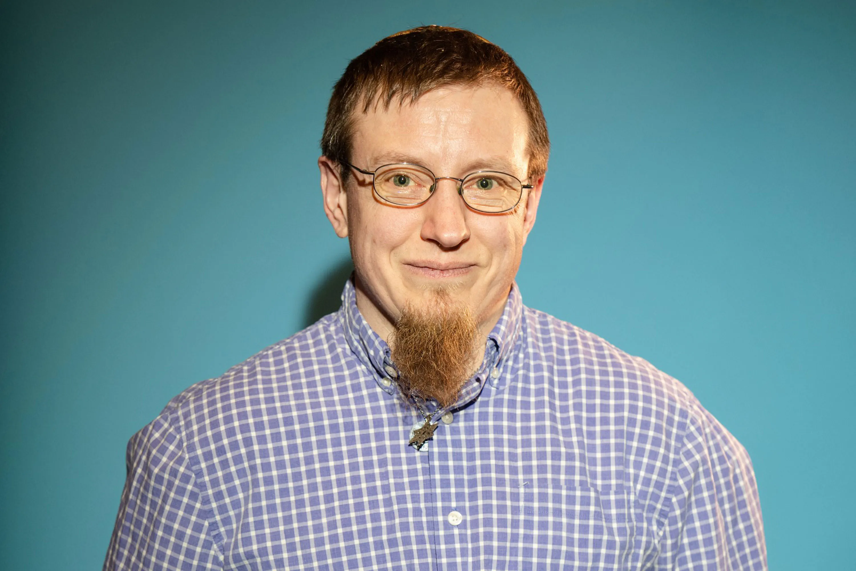 Portrait of Lis Regula. He has short brown hair, glasses, and a long, pointed beard is smiling softly. He wears a light purple and white checked shirt and stands in front of a plain blue background.
