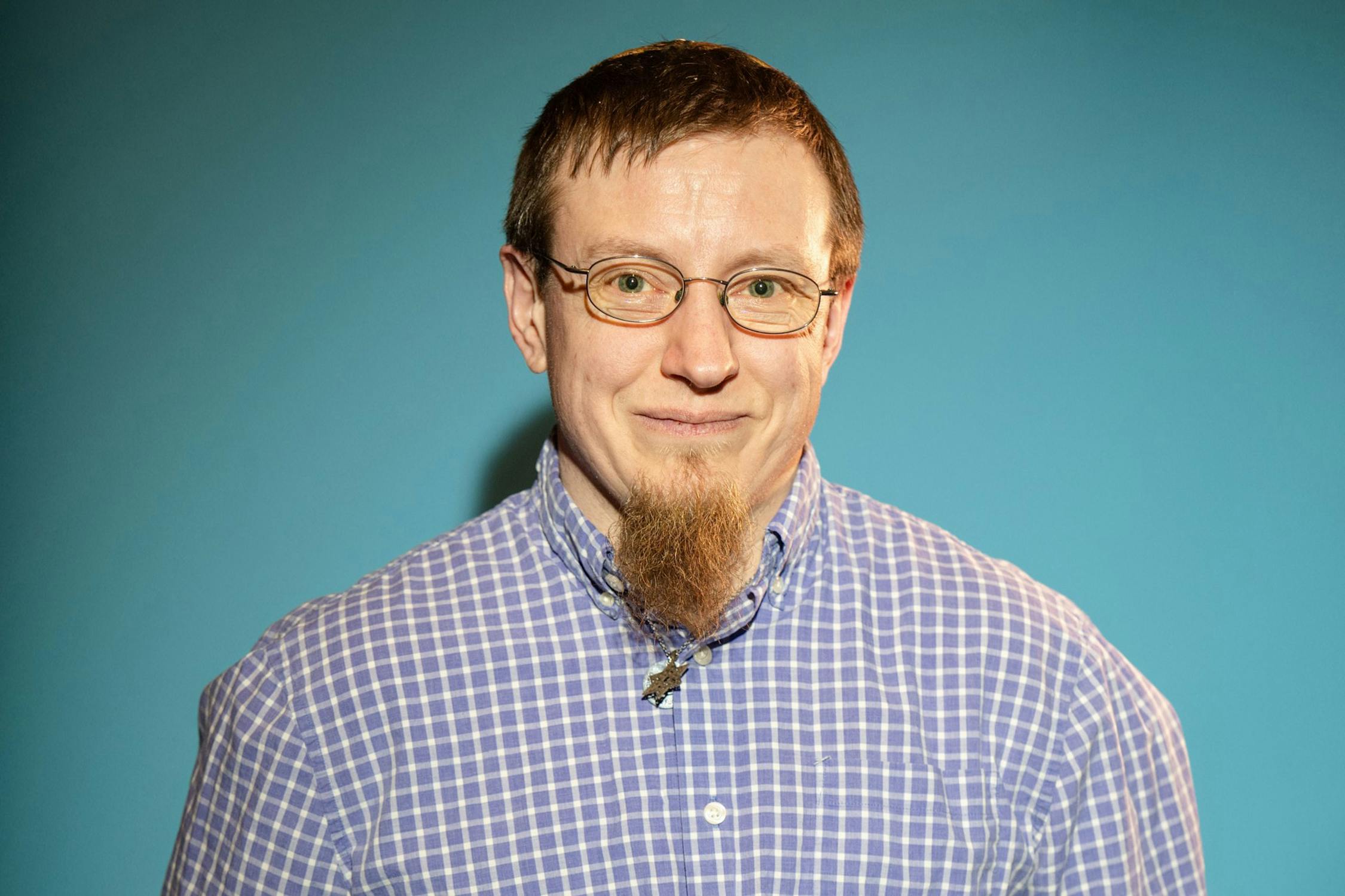 Portrait of Lis Regula. He has short brown hair, glasses, and a long, pointed beard is smiling softly. He wears a light purple and white checked shirt and stands in front of a plain blue background.