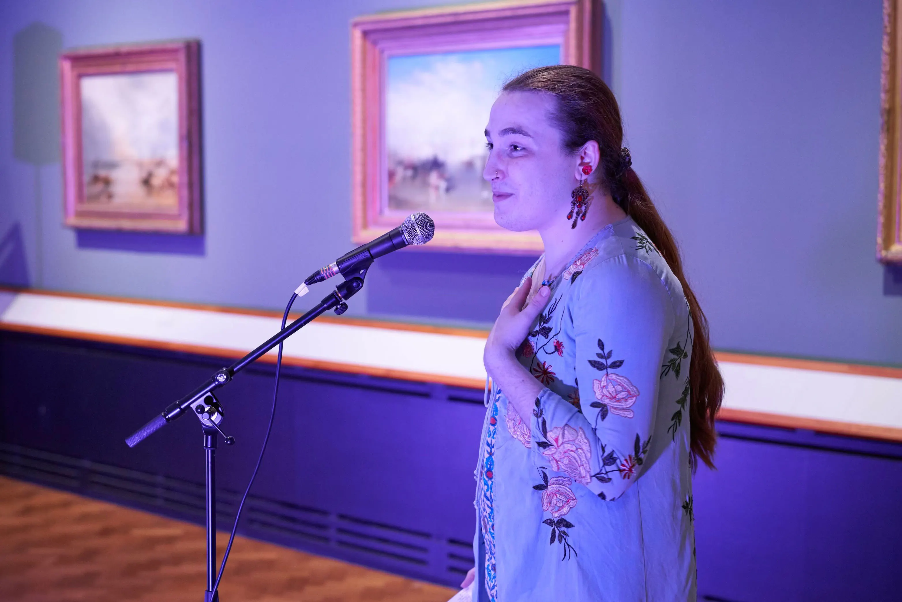 Cas Bardberr, standing at a mic. She has long brown hair and is wearing a light blue floral top. They rest a hand on their chest and speak, with several framed paintings hanging on a blue wall behind them. The scene is softly lit.