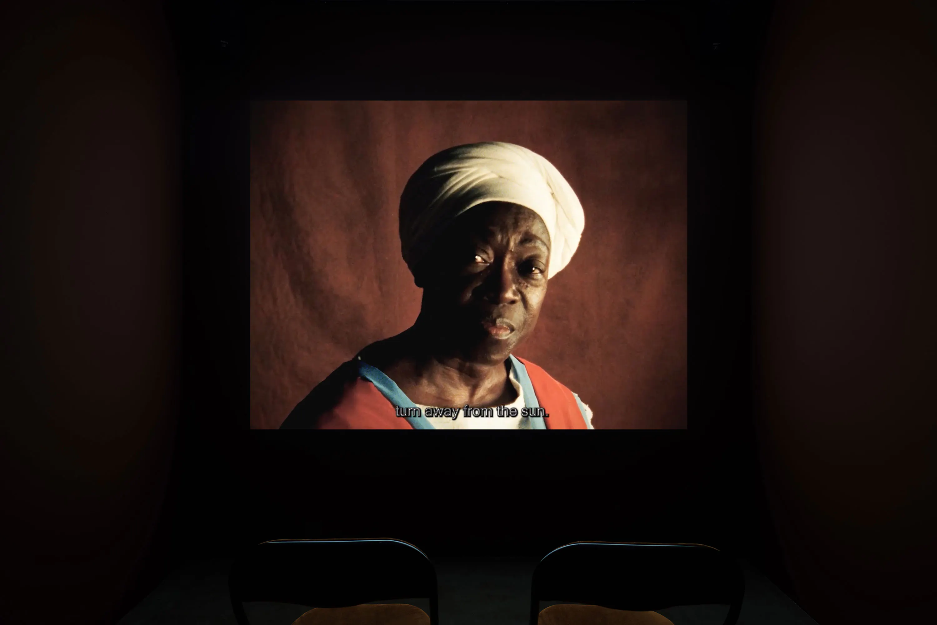 A dimly lit room with two empty chairs faces a projected film image of an older black woman in a white head wrap and red clothing. The screen shows English subtitles reading, “turn away from the sun,” against a reddish background.