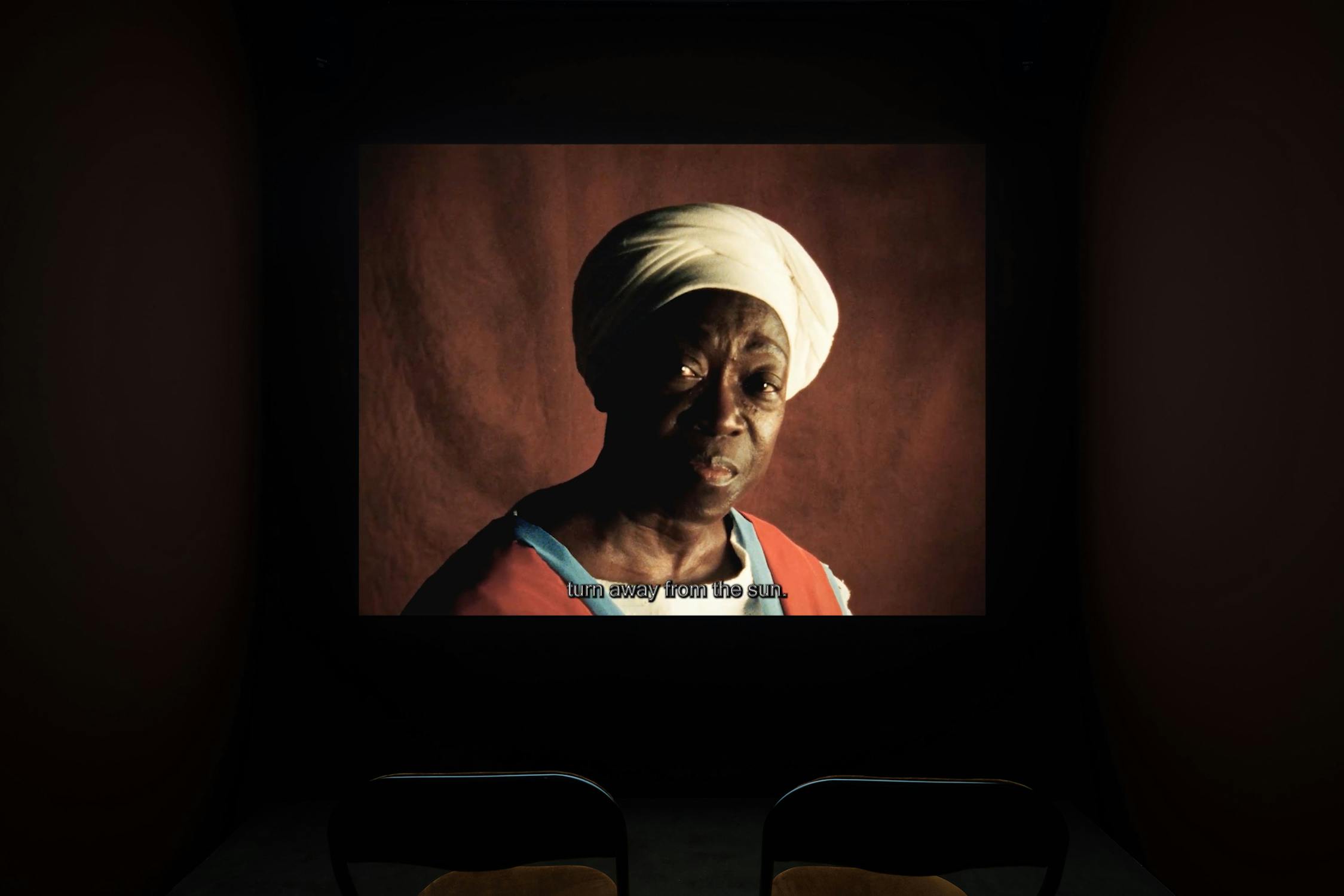 A dimly lit room with two empty chairs faces a projected film image of an older black woman in a white head wrap and red clothing. The screen shows English subtitles reading, “turn away from the sun,” against a reddish background.