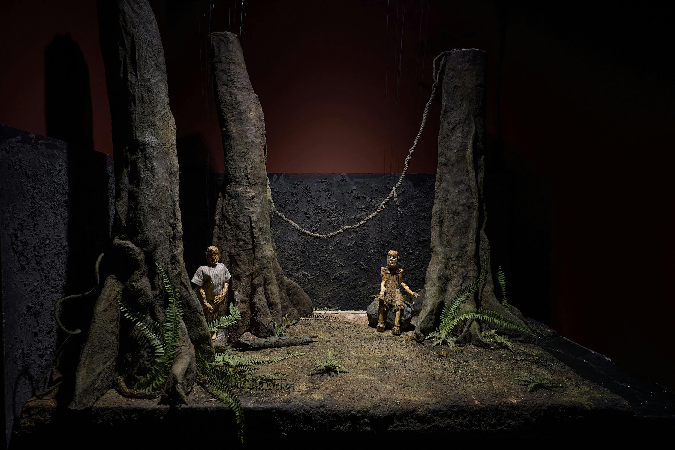 A dimly lit scene features two puppets sitting on a forest floor surrounded by tall, bare tree trunks and ferns. One puppet sits near the base of a tree, while the other is on a wooden stump. The background is dark and textured, evoking a mysterious forest.