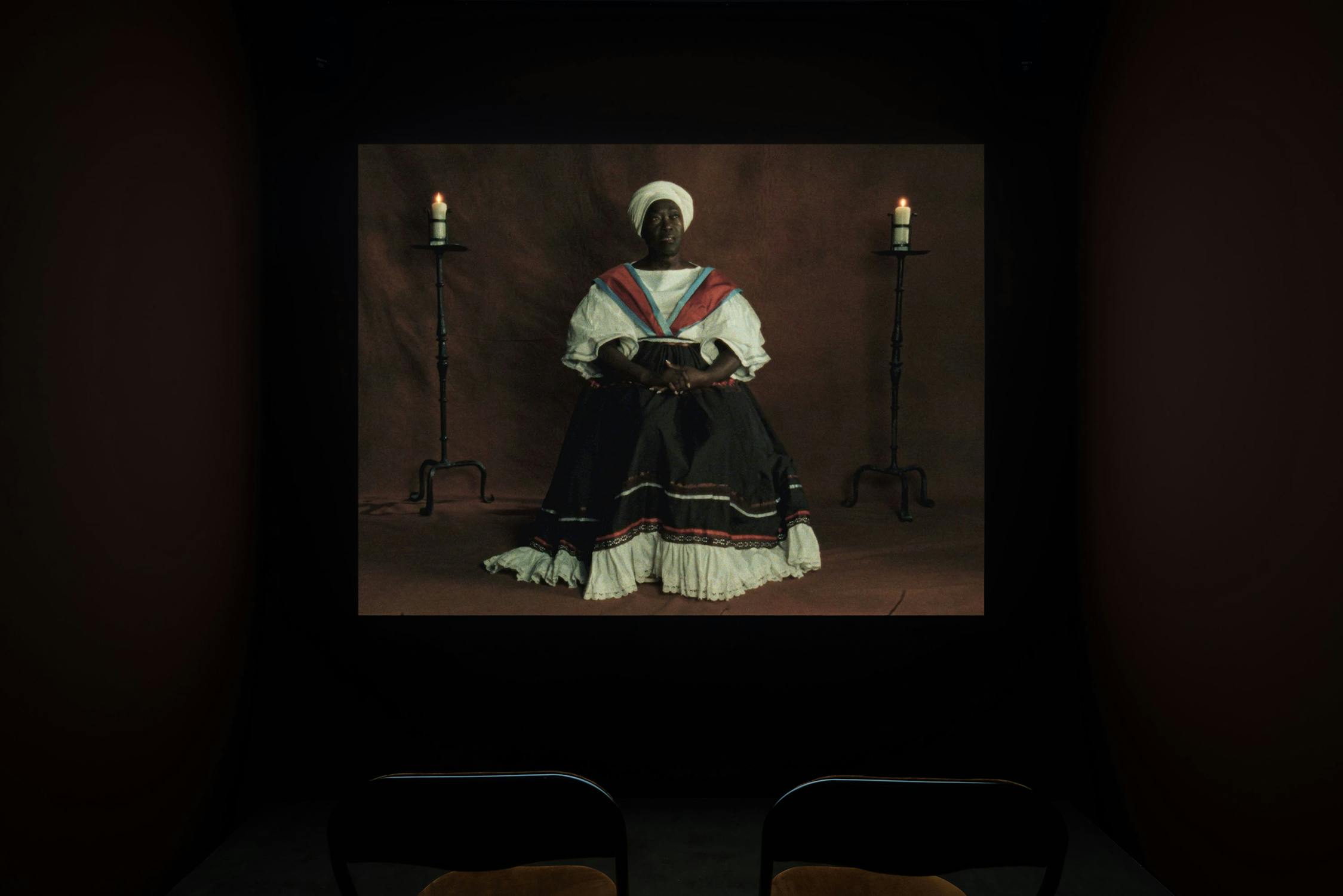 A dimly lit room shows a projected image of a Black woman in historical dress, seated with hands in her lap. She wears a white bonnet, layered black dress with red, blue, and white accents, and sits between two tall lit candles on stands. Two empty chairs face the projection.