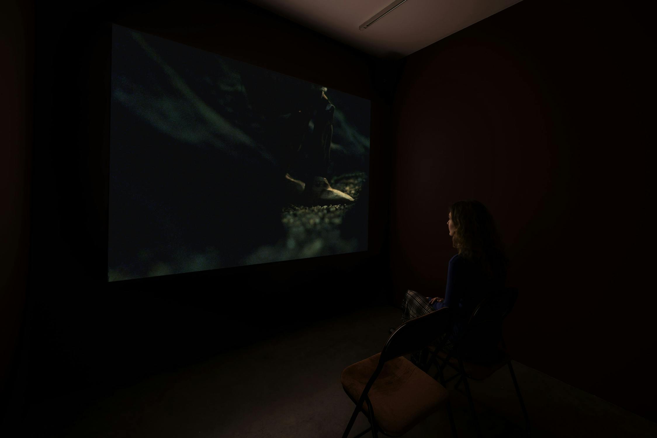 A person with long hair sits alone in a dark room on a folding chair, watching a dimly lit film projected on a large wall screen. The image on the screen shows a close-up of feet in low light. The room is sparsely furnished and shadowy.