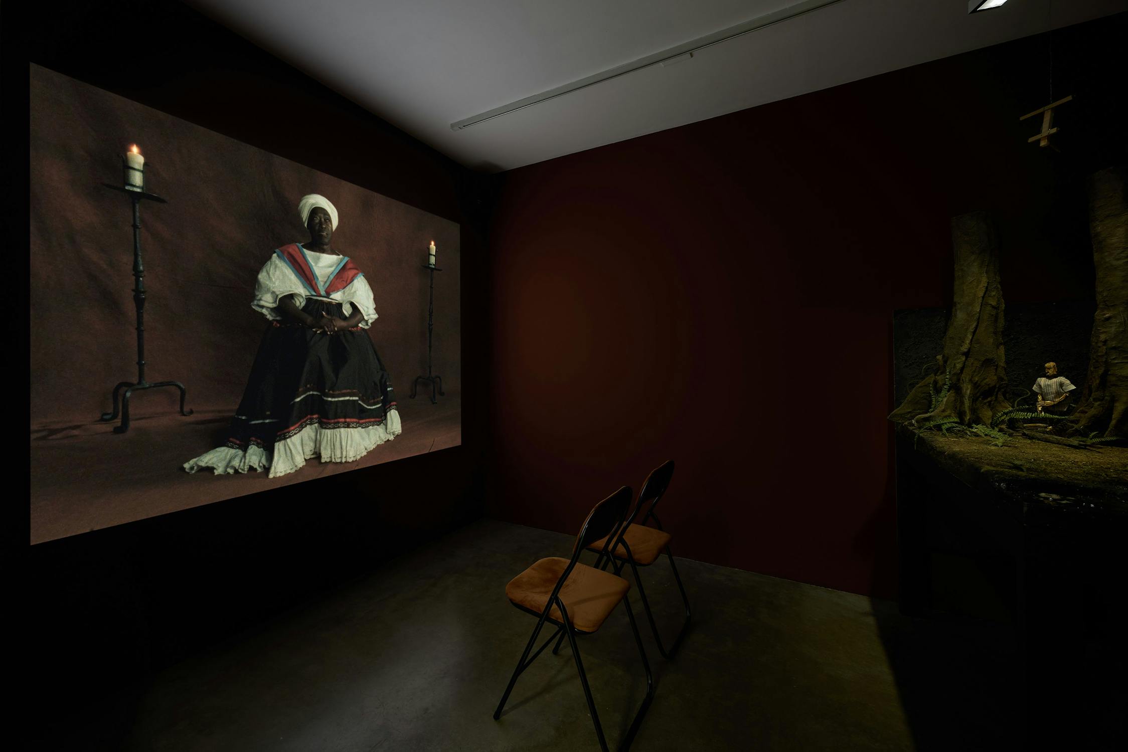 A dimly lit gallery room features two folding chairs facing a large projected portrait of a seated woman in a historic dress, flanked by candlesticks. To the right, a small diorama depicts a figure in a forest-like setting. The walls are dark brown, creating an intimate atmosphere.