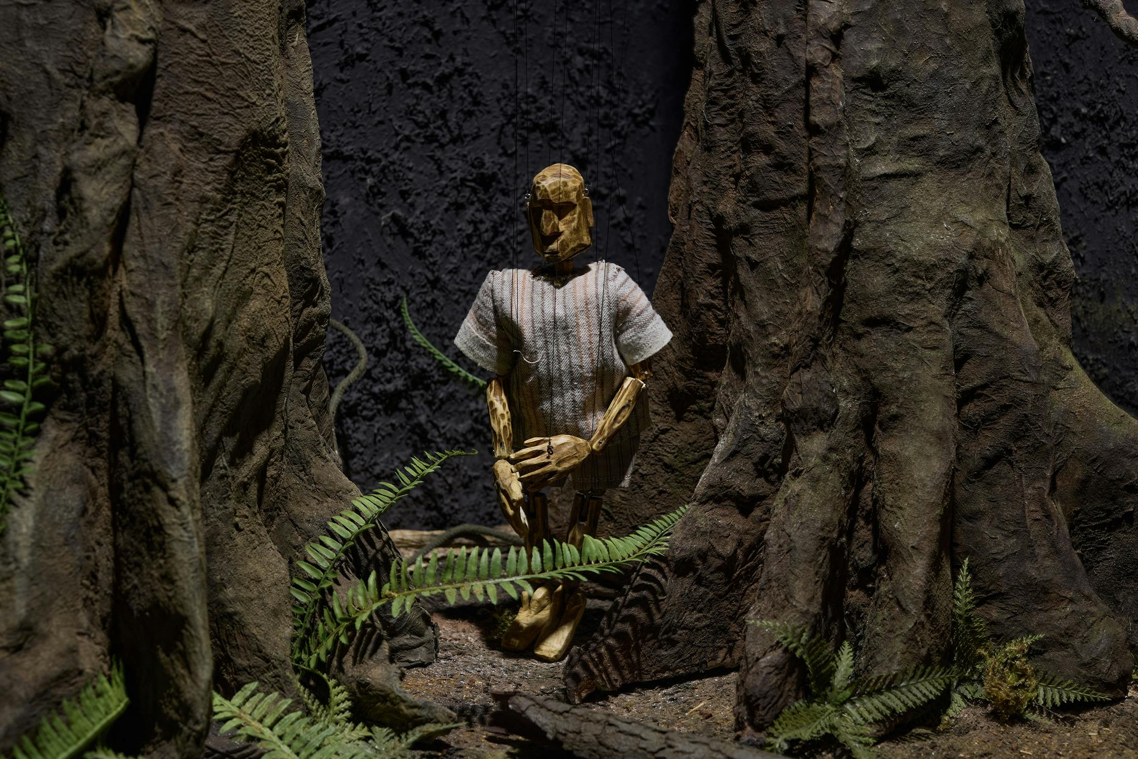 A wooden puppet wearing a striped short-sleeve shirt kneels on the forest floor between large tree trunks. Ferns grow around the puppet and trees, and the rough dark backdrop suggests a dense, shadowy woodland scene.