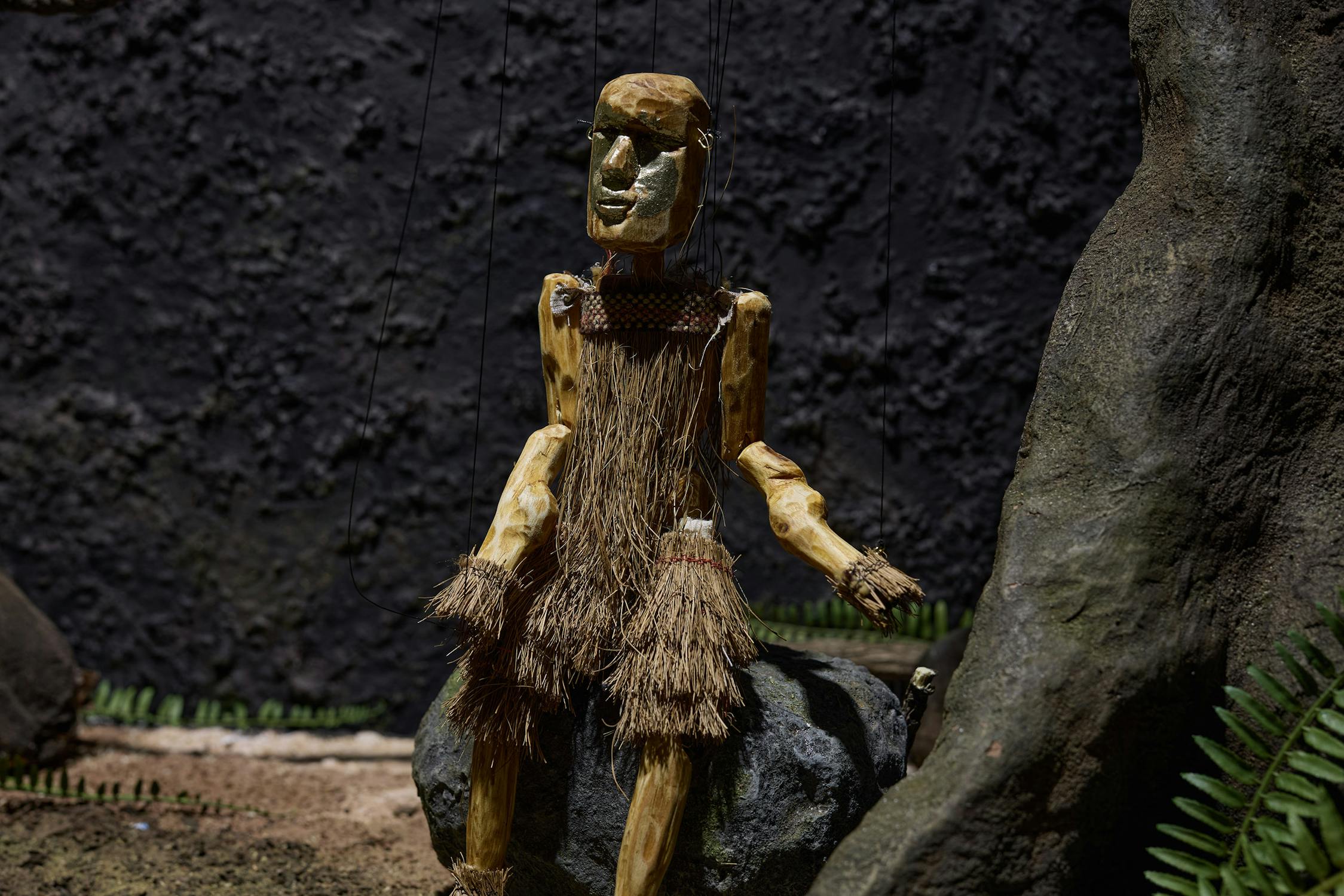 A wooden marionette figure with a grass skirt sits on a rock. The puppet has a carved face, string joints, and straw details on its arms and torso. Ferns and a large tree trunk are nearby, with a dark, textured backdrop behind them.