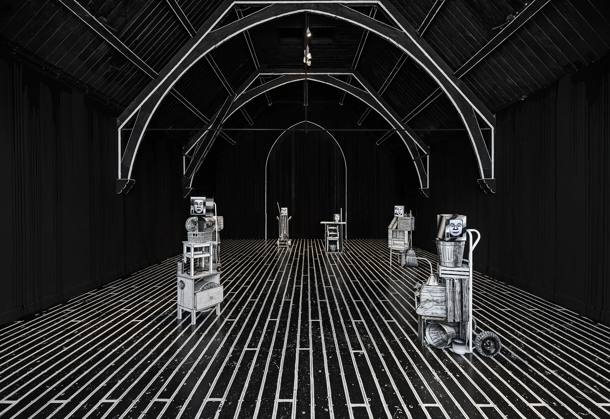 A black room with a vaulted ceiling features five metal sculptures on wheeled bases, spaced apart. White lines on the floor and arches highlight the geometric perspective, whilst the sculptures resemble assembled household objects, creating an industrial, surreal atmosphere.