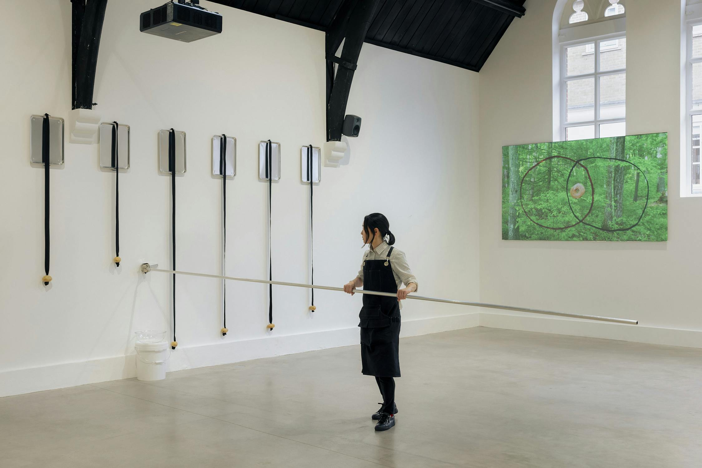 Aki Sasamoto, in a black apron, stands in a gallery holding a long silver pole. On the white wall, seven rectangular sheet pans with elastic cords hang vertically. A green artwork with circular patterns hangs on the right wall beneath tall arched windows.