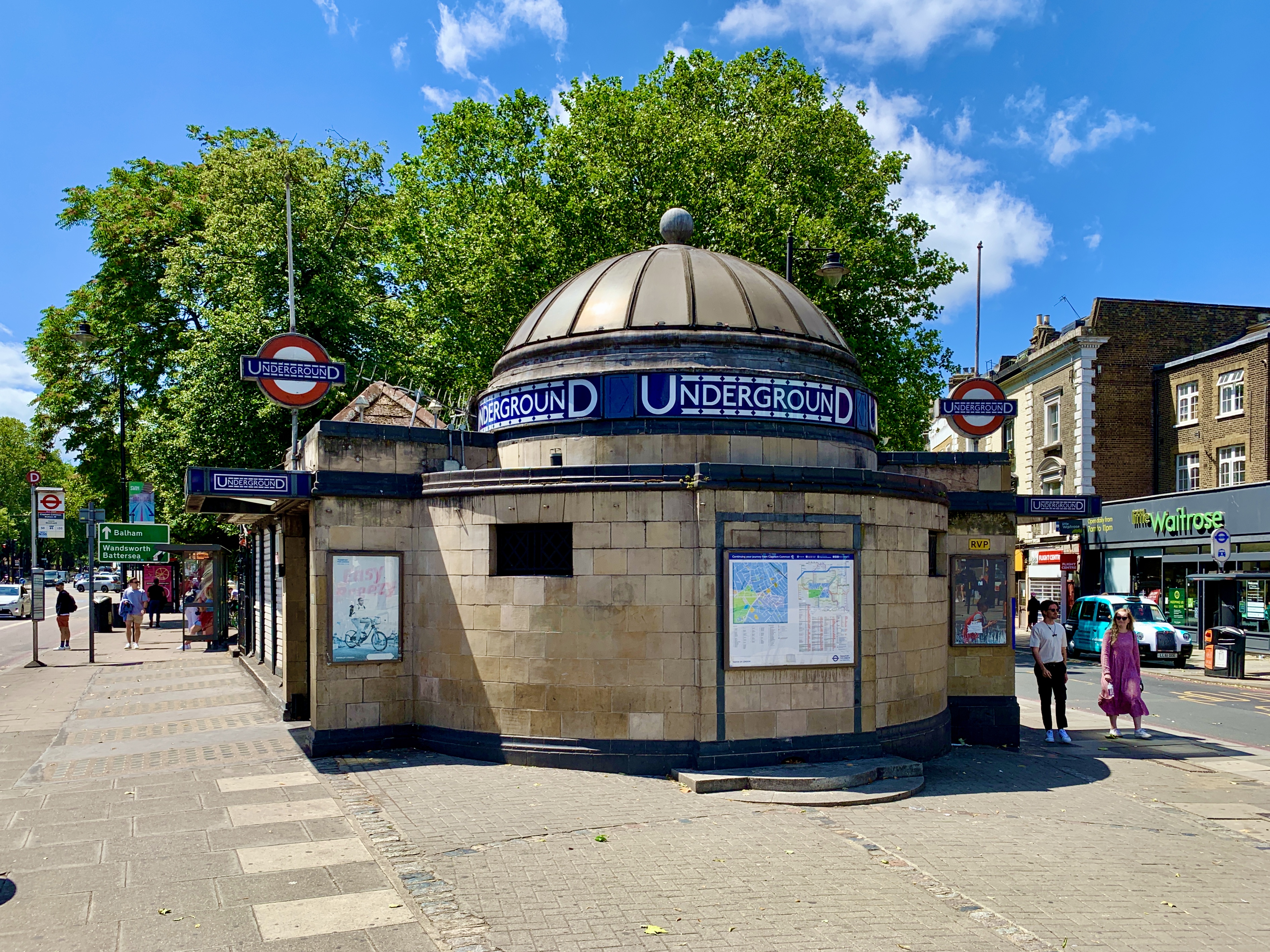 Circular stone entrance to a London Underground station, with a domed roof and four red-blue Underground signs. Several people stand outside, some looking at a map. Trees and shops are visible in the background on a bright day.