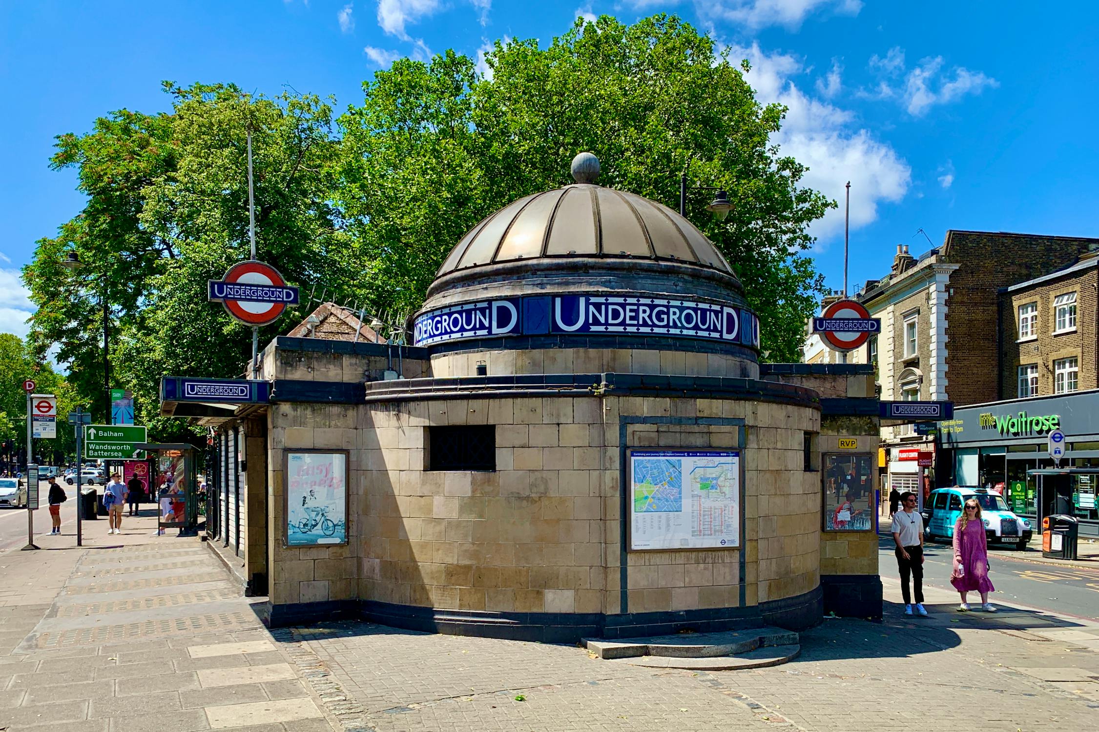 Circular stone entrance to a London Underground station, with a domed roof and four red-blue Underground signs. Several people stand outside, some looking at a map. Trees and shops are visible in the background on a bright day.